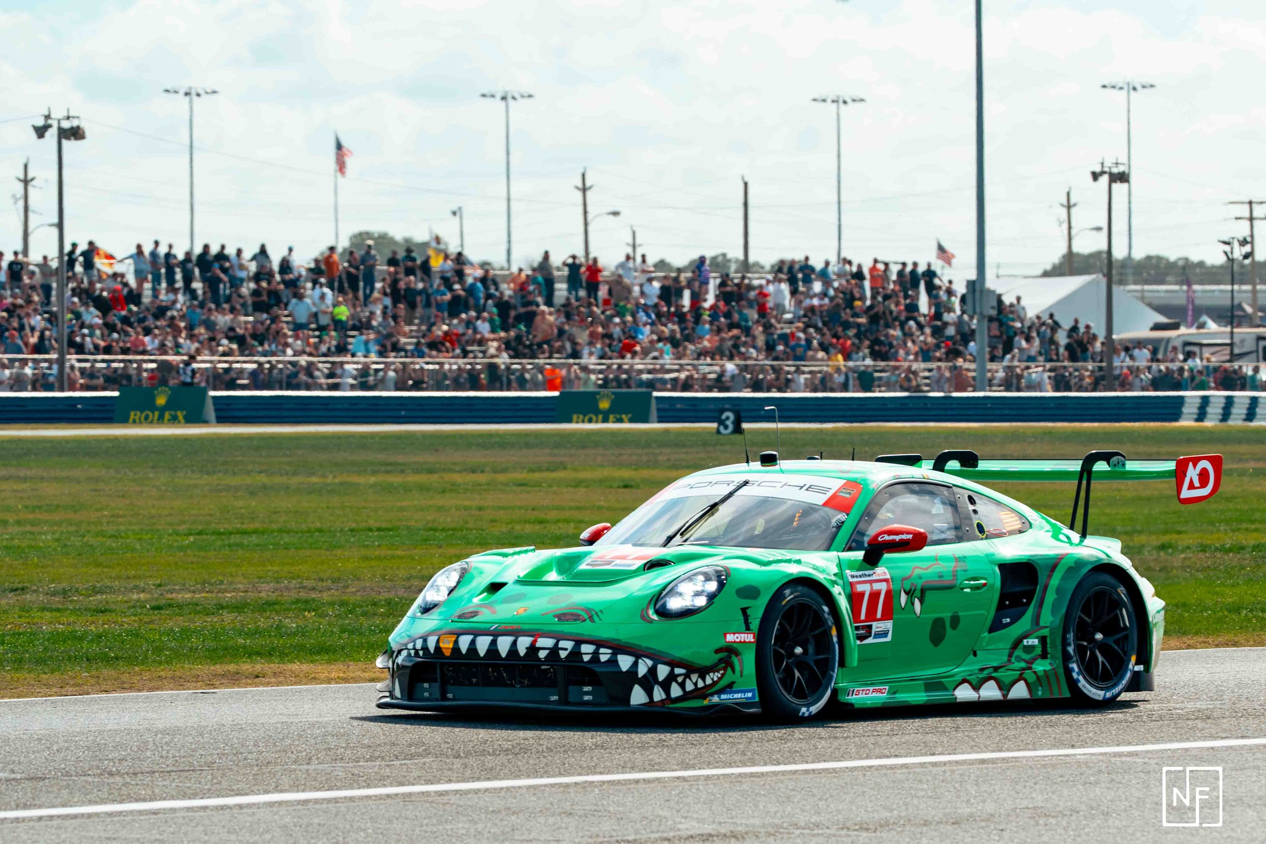 Green race car with a dragon face design racing on a track, crowd and grandstands in the background.