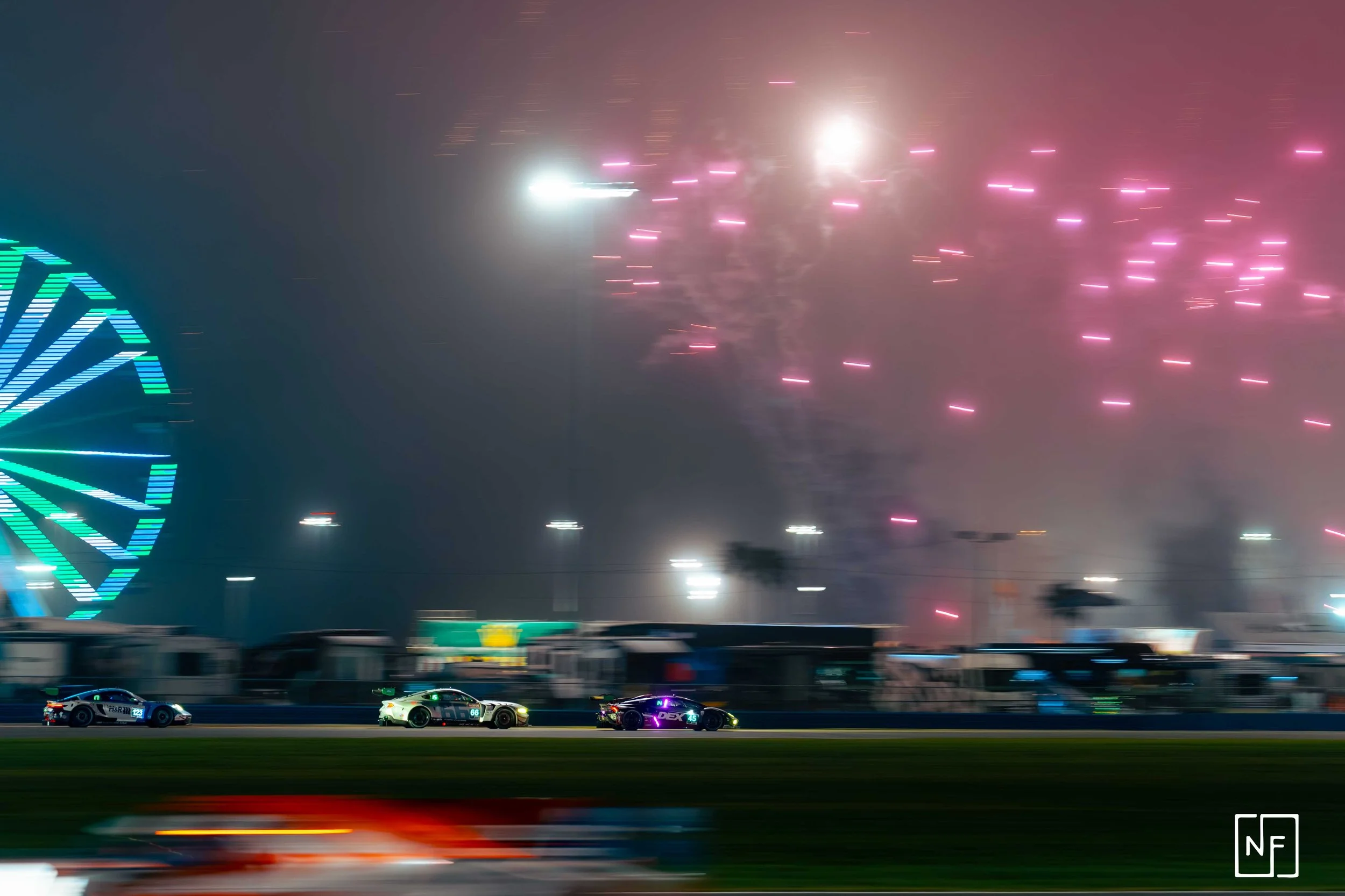 Nighttime scene of a race track with race cars speeding, brightly lit ferris wheel to the left, and colorful fireworks in the sky.