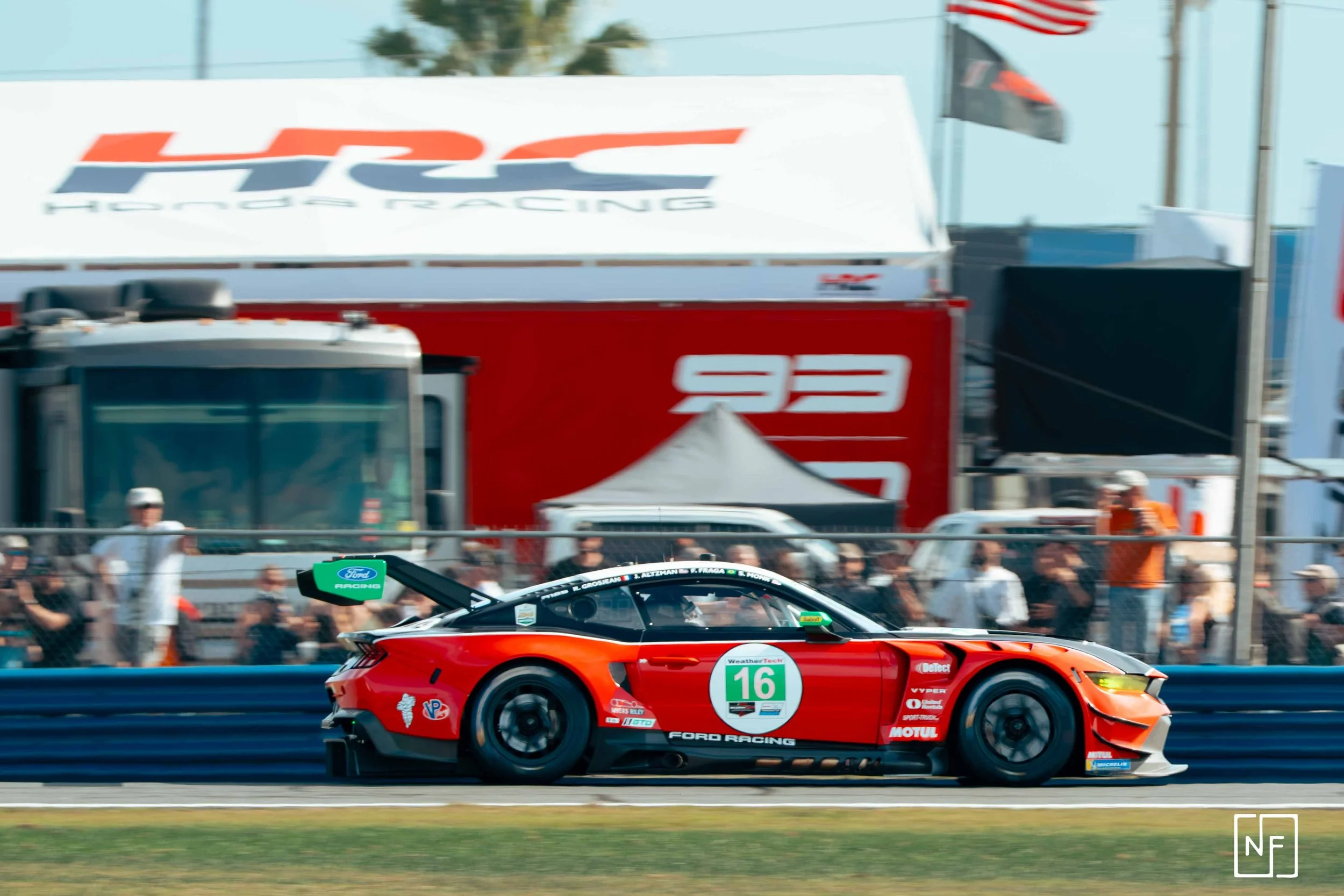 A red Ford race car, number 16, moving on a race track with spectators watching behind a fence in the background.
