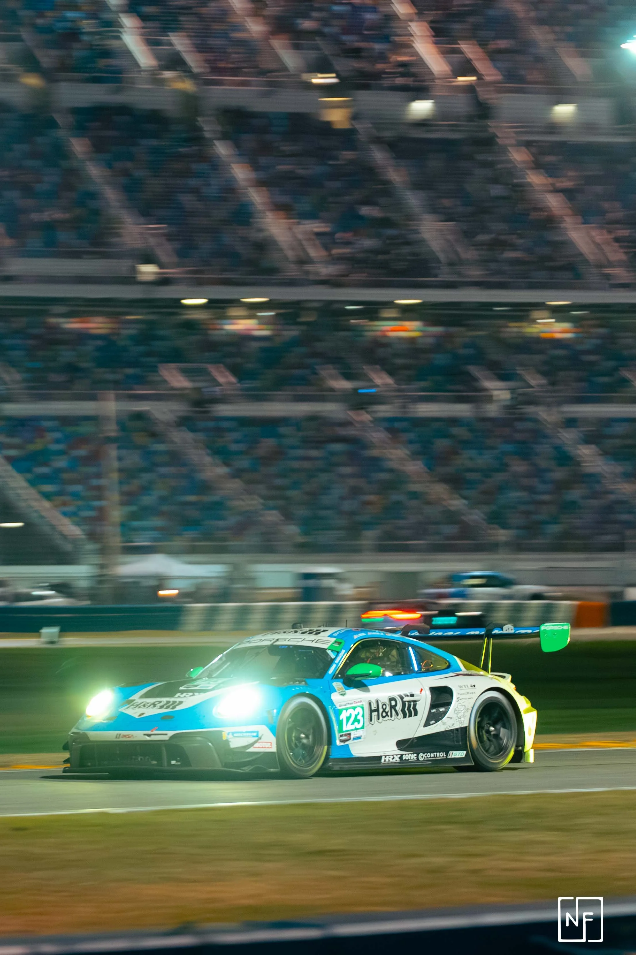 Race car on a track at night with motion blur. The car has a blue, white, and green color scheme with the number 123 and sponsors such as H&R and Porsche. The background shows blurred spectators in the stands.