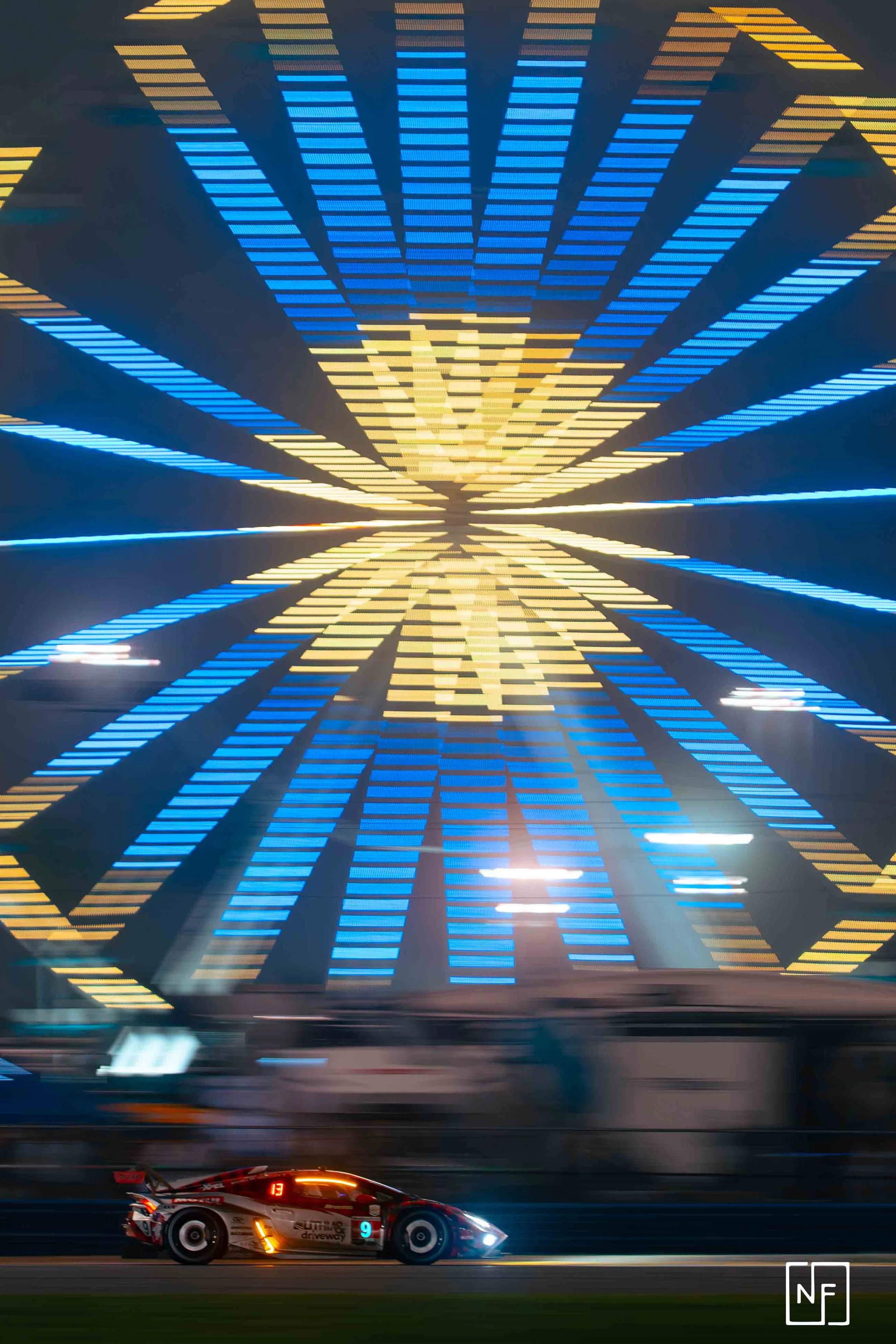 Long exposure photo of a race car on a track with a brightly illuminated Ferris wheel in the background.