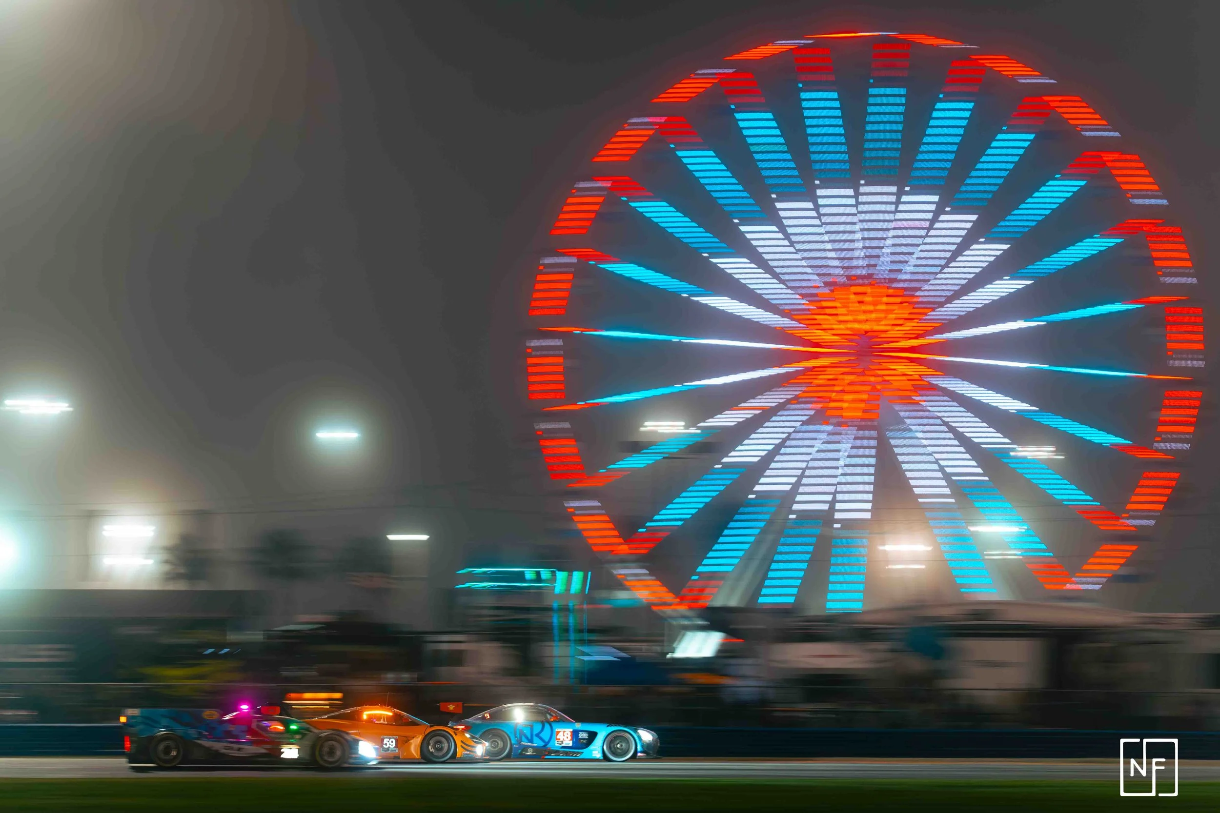 Nighttime scene at a racetrack with three racing cars in the foreground, illuminated by colorful lights, and a large, brightly lit Ferris wheel in the background.