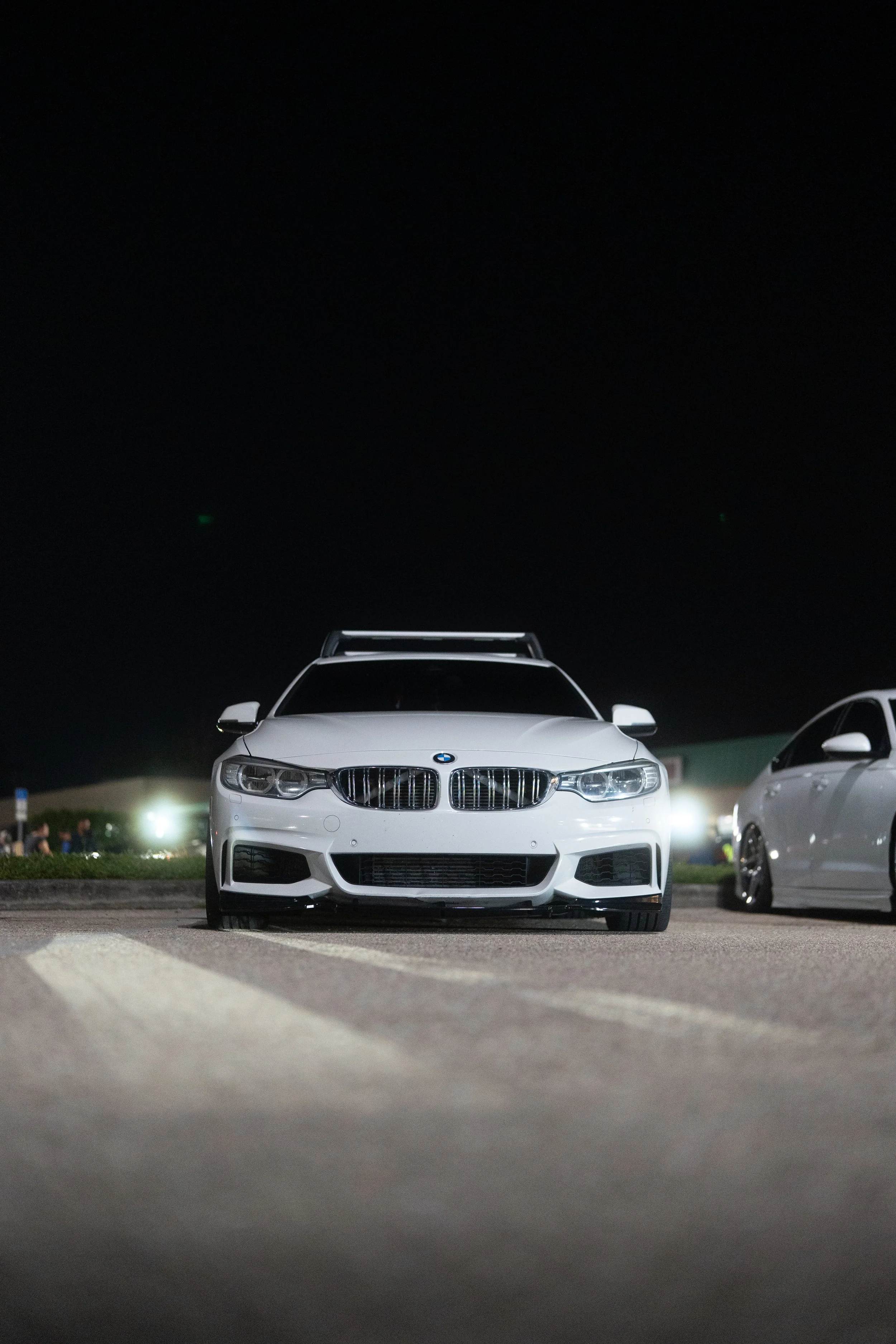 White BMW car parked in a parking lot at night with other cars and blurred lights in the background.