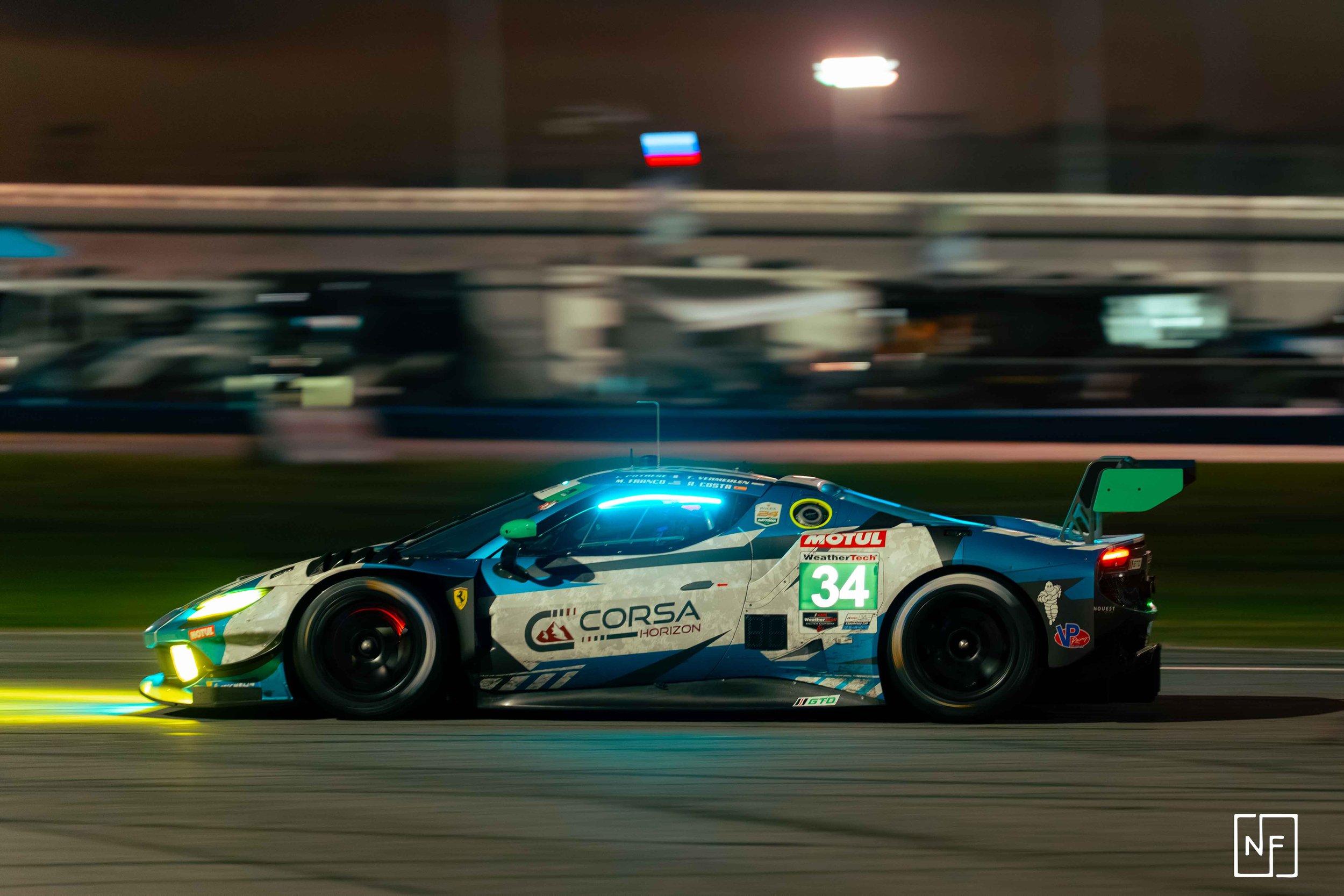 A race car on the track at night, illuminated by game lights, with sponsorship logos, a large rear wing, and the number 34 on the side.