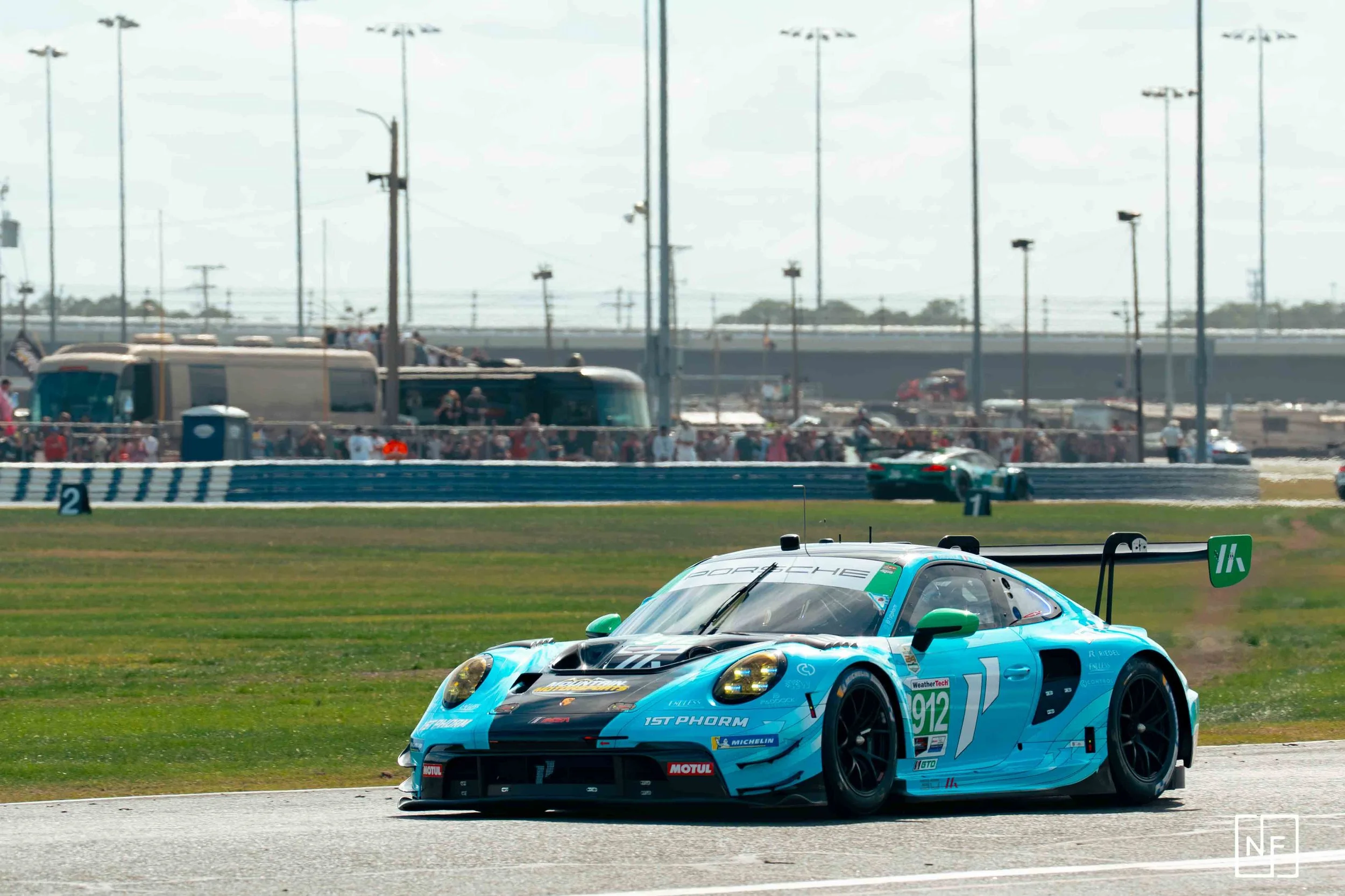 A blue Porsche race car on a racetrack with a crowd and vehicles in the background.