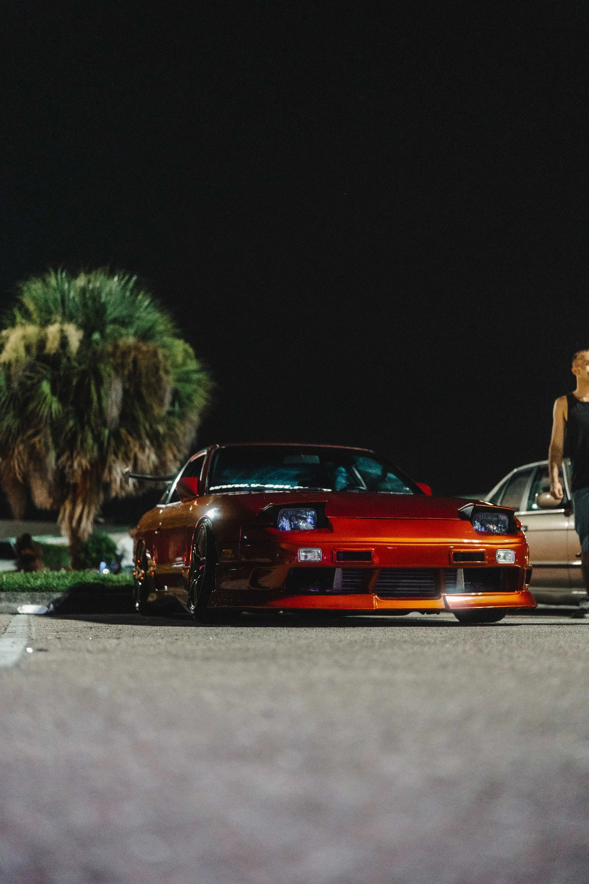 Red sports car parked in a lot at night, with a palm tree and a person on the right side of the image.
