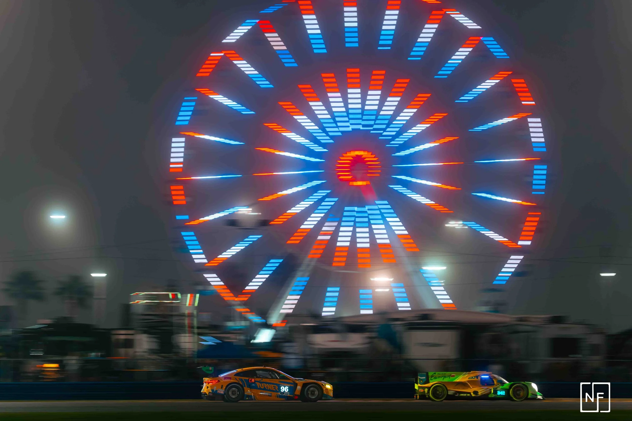 Nighttime scene of a Ferris wheel with colorful red, white, and blue lights. Below, two race cars speed on a track, with one car front and center and the other slightly behind, both brightly lit.