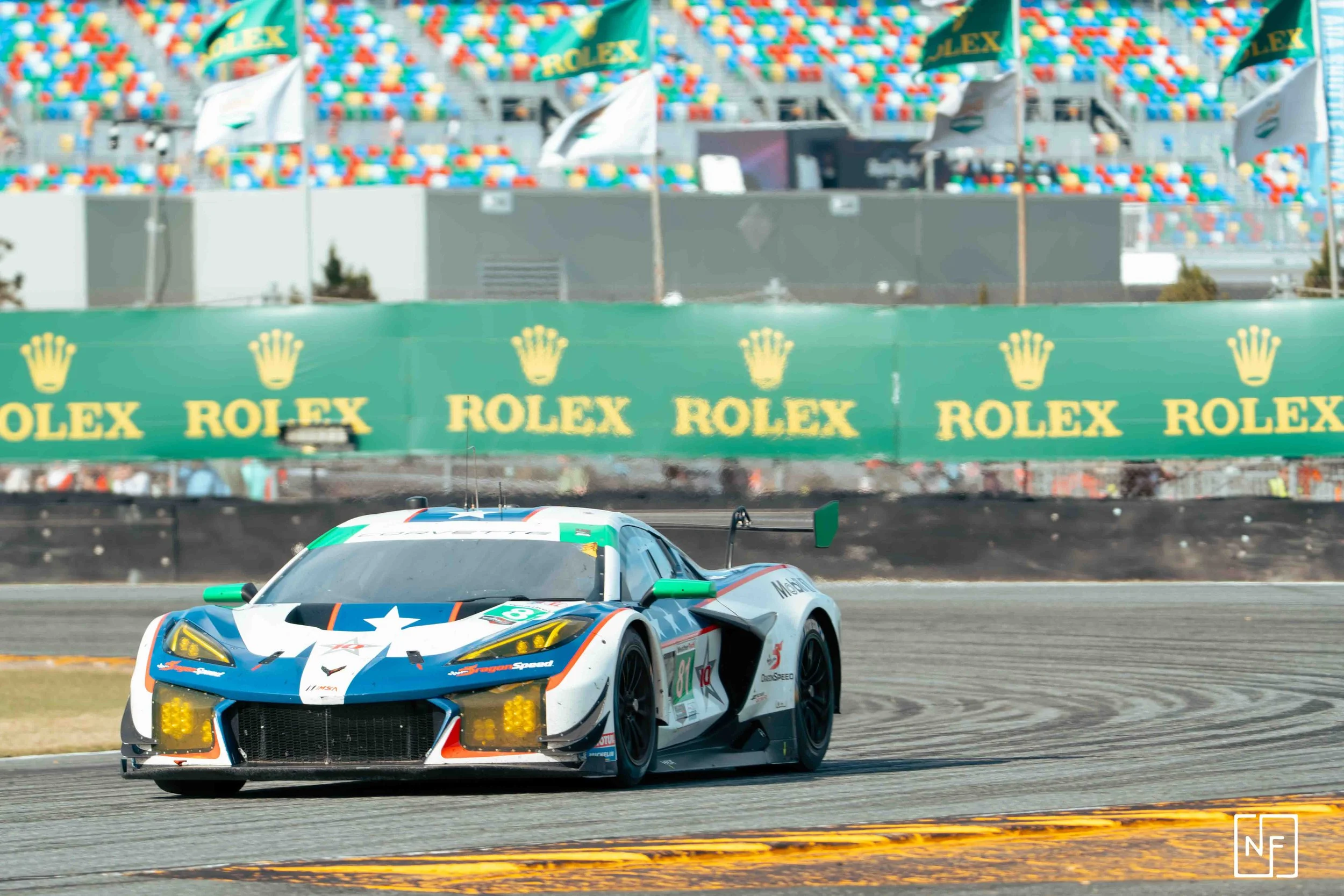 A race car with star and stripe decals on a track, with green and yellow Rolex advertising banners and colorful seats in the background.