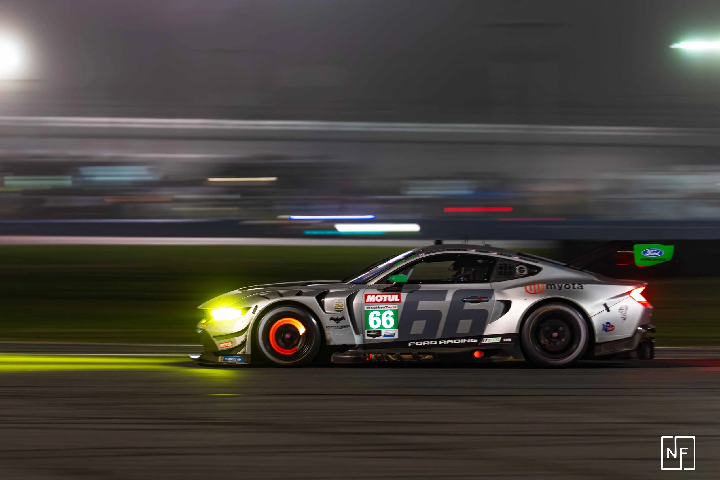 Race car speeding on track at night with motion blur, green and yellow headlamps, racing decals, number 66, Ford Racing logo, and sponsorship stickers.