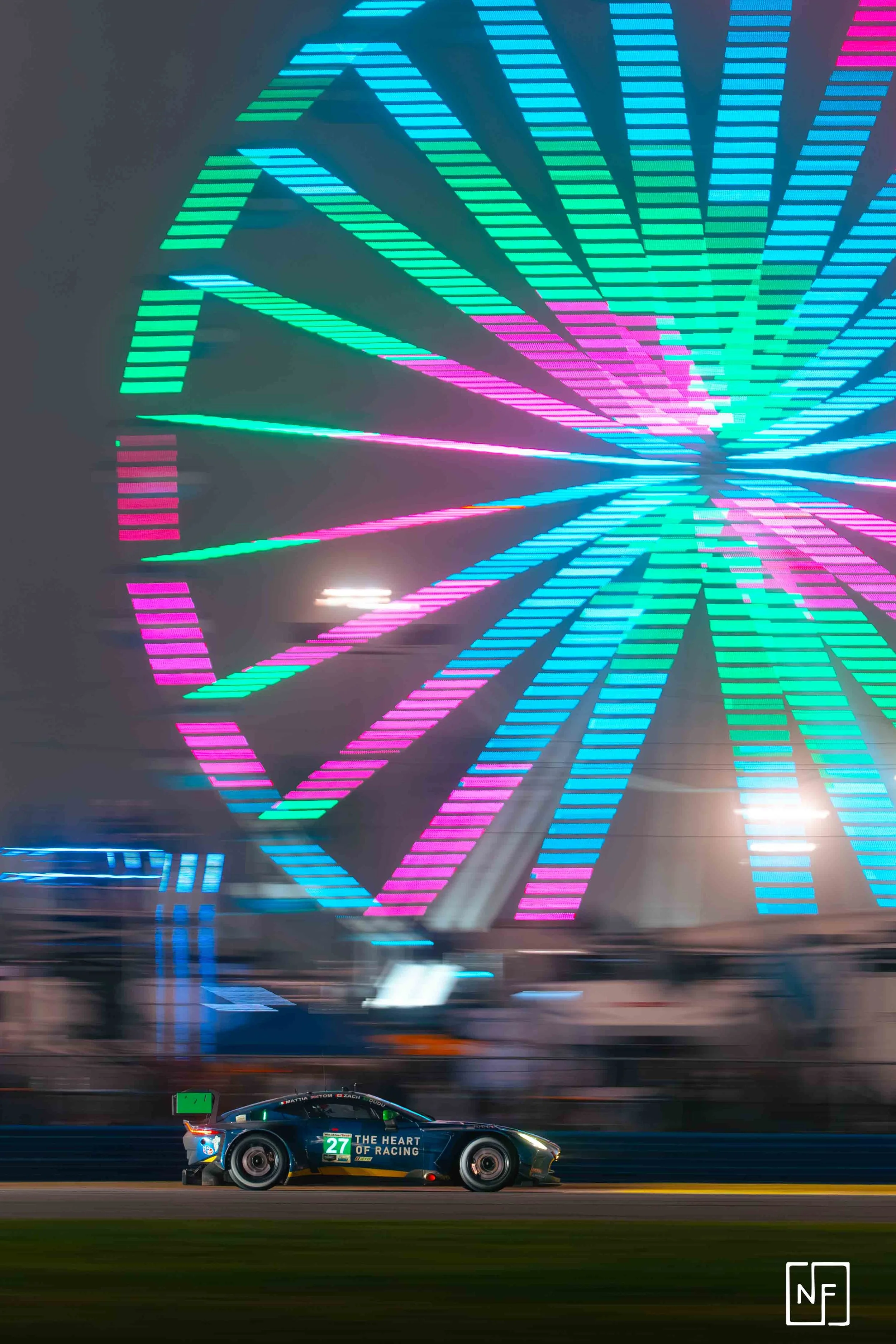 A race car on the track with a brightly lit ferris wheel in the background at night.