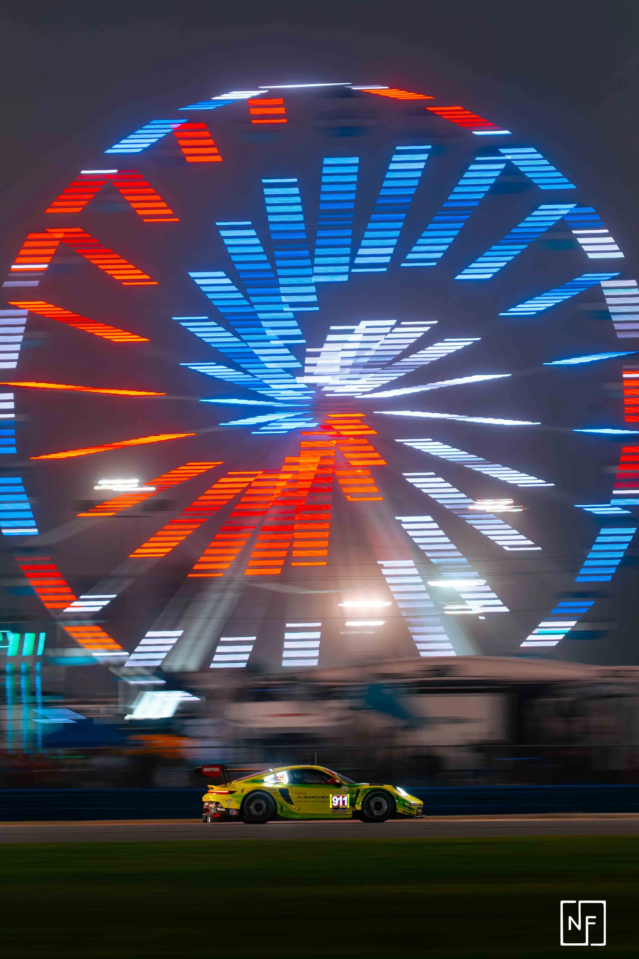A yellow race car driving on a track at night with a large, colorful Ferris wheel illuminated in the background.