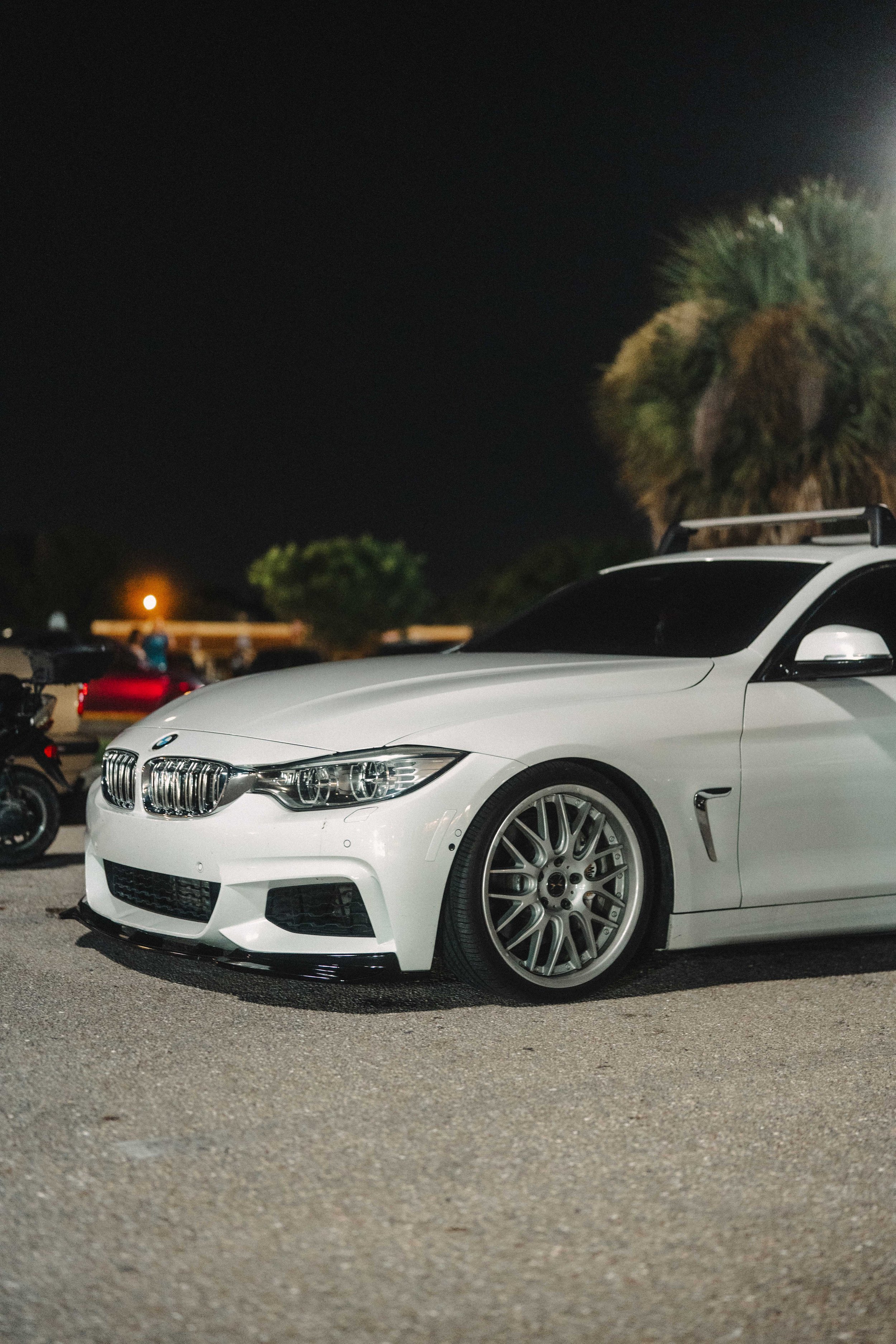 A white BMW sports car parked at night in a lot, with trees and other cars visible in the background.