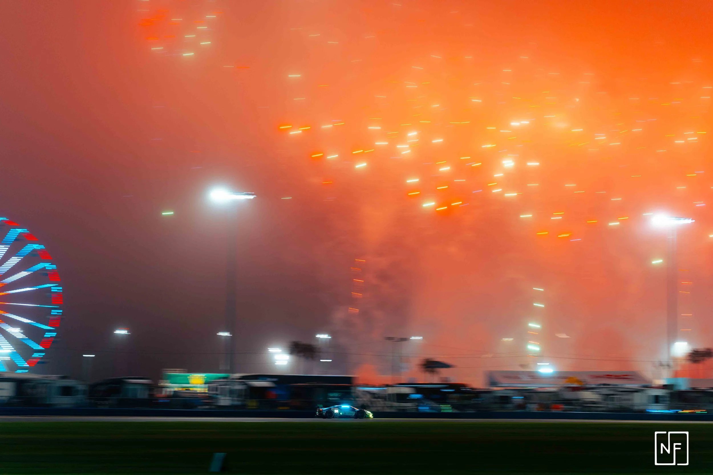 Fireworks explode in the night sky over a racetrack, with a racing car on the track and a large ferris wheel illuminated on the left side.