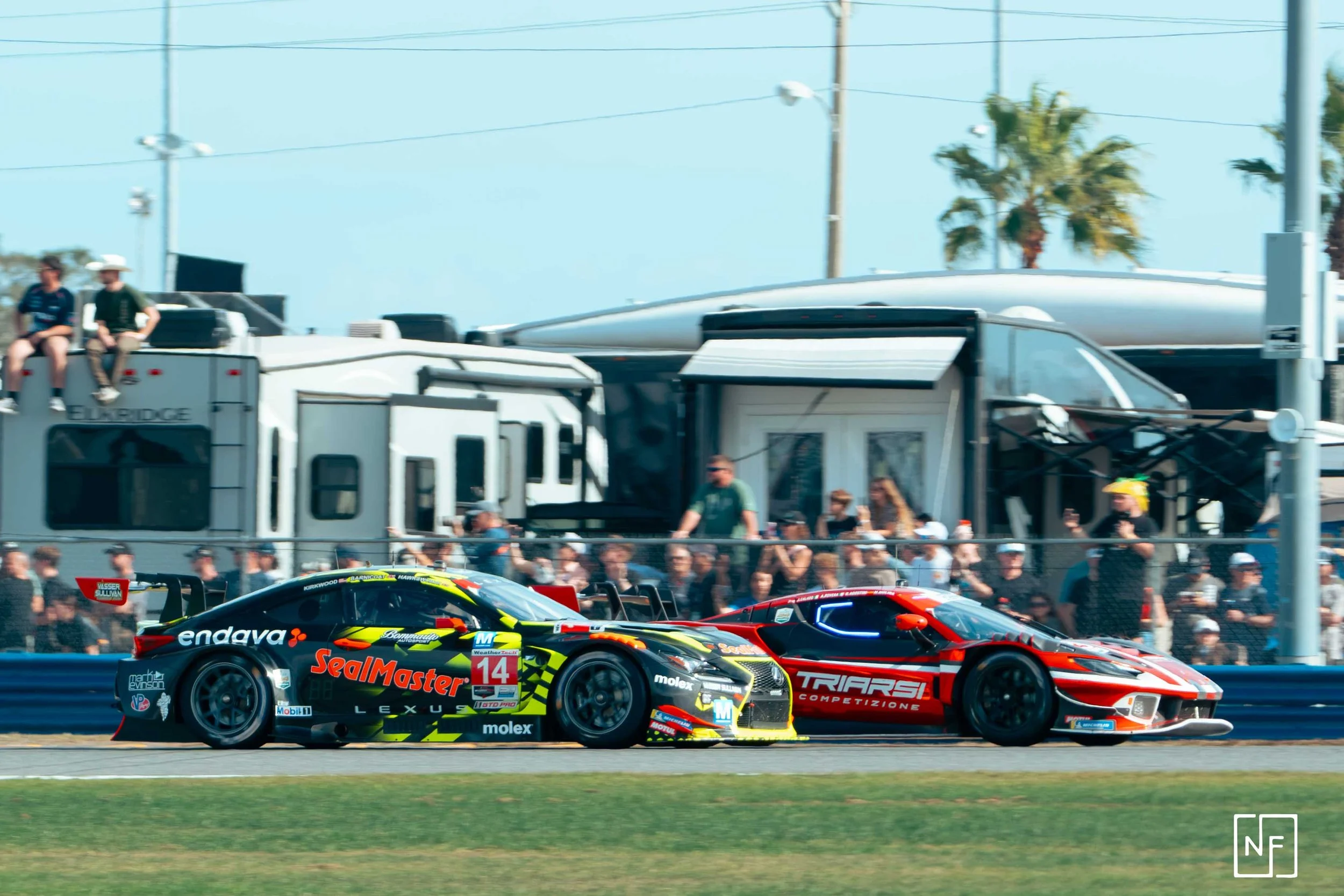 Two race cars speeding on a racetrack, with a crowd of spectators and RVs in the background.