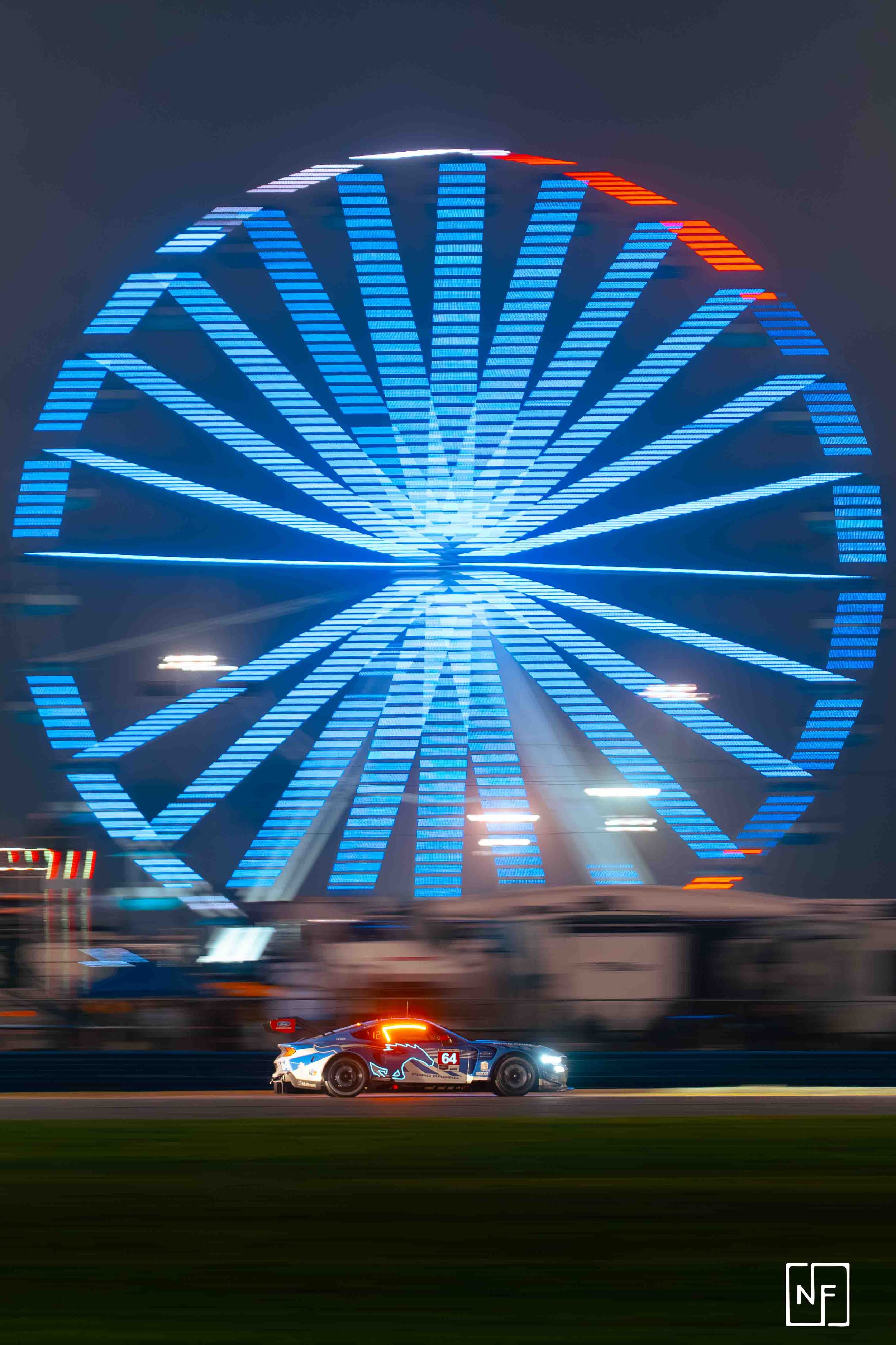 A race car moving at night in front of a brightly lit Ferris wheel with blue and red lights.