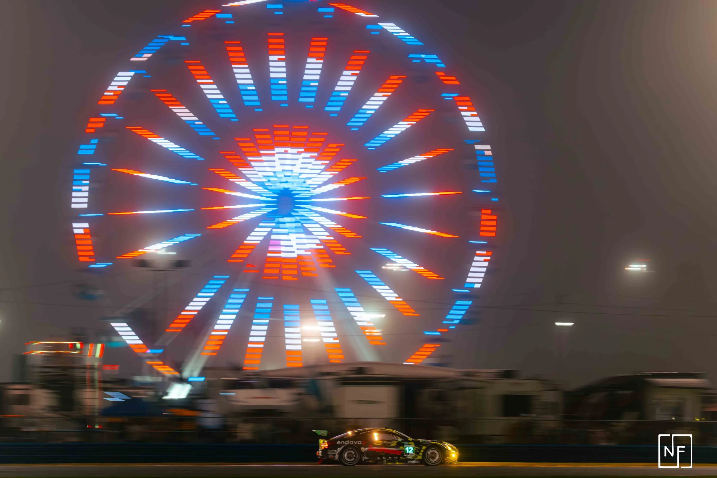 A racing car driving past a large illuminated Ferris wheel at night, with the wheel displaying bright red, blue, and white lights in a circular pattern.