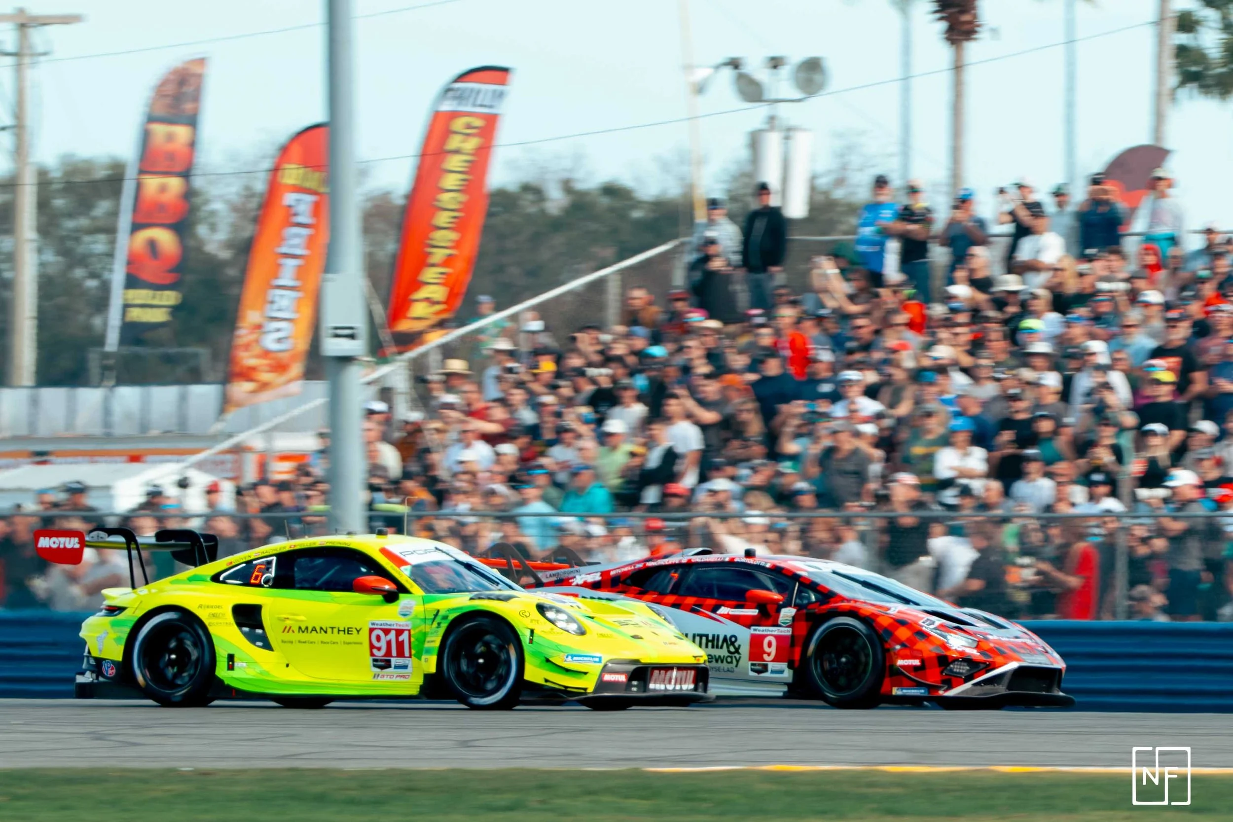 Two race cars, yellow and red, racing on a track with a large crowd of spectators in the background, colorful flags, and a clear sky.
