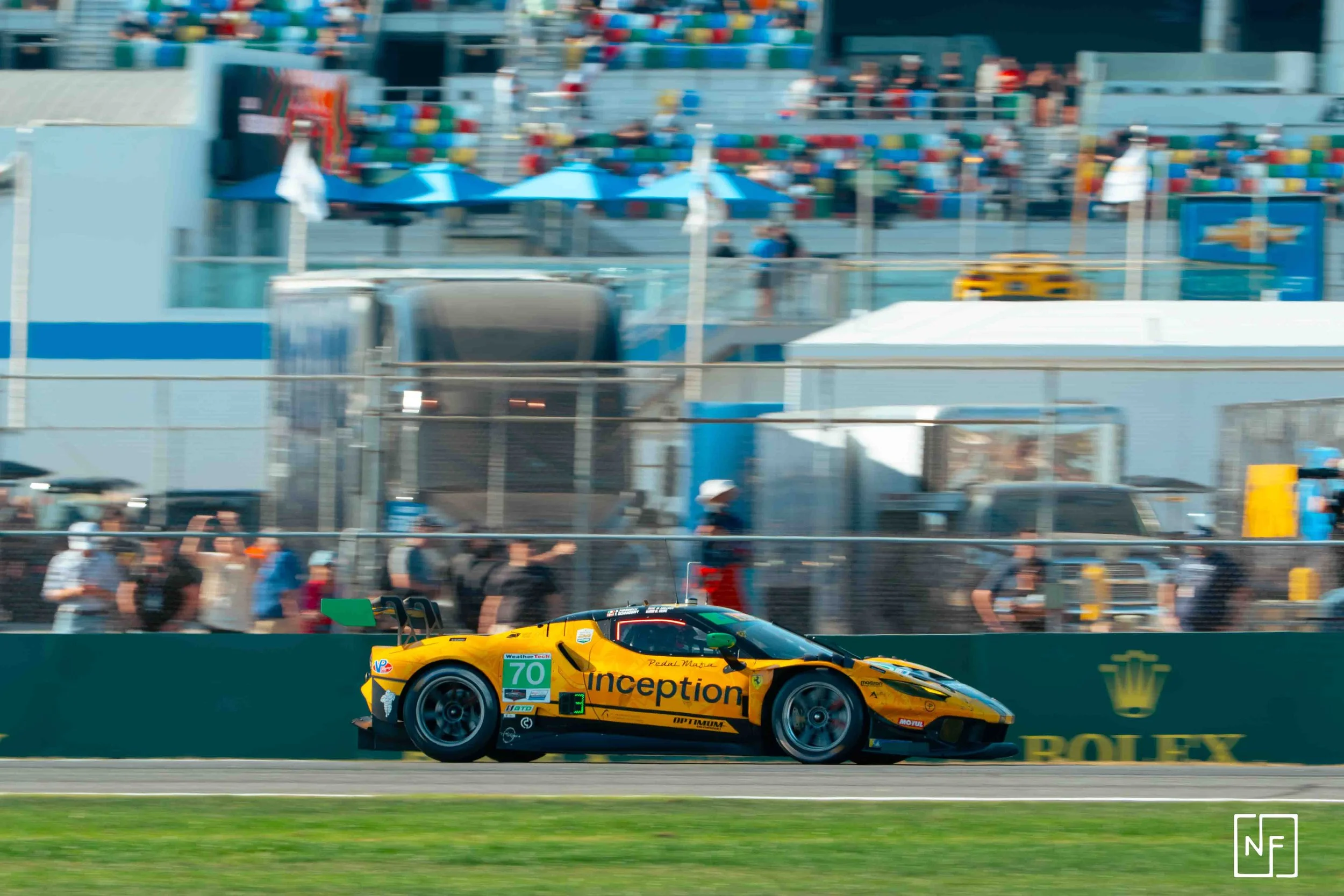 Race car on a track with spectators and team members in the background, Rolex signage on the barrier.