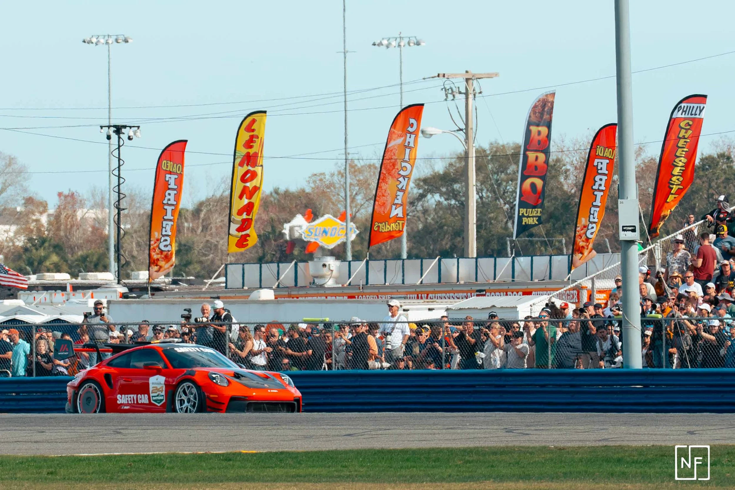 A red safety race car on a racetrack with a crowd of spectators behind a fence and colorful flags waving in the background.