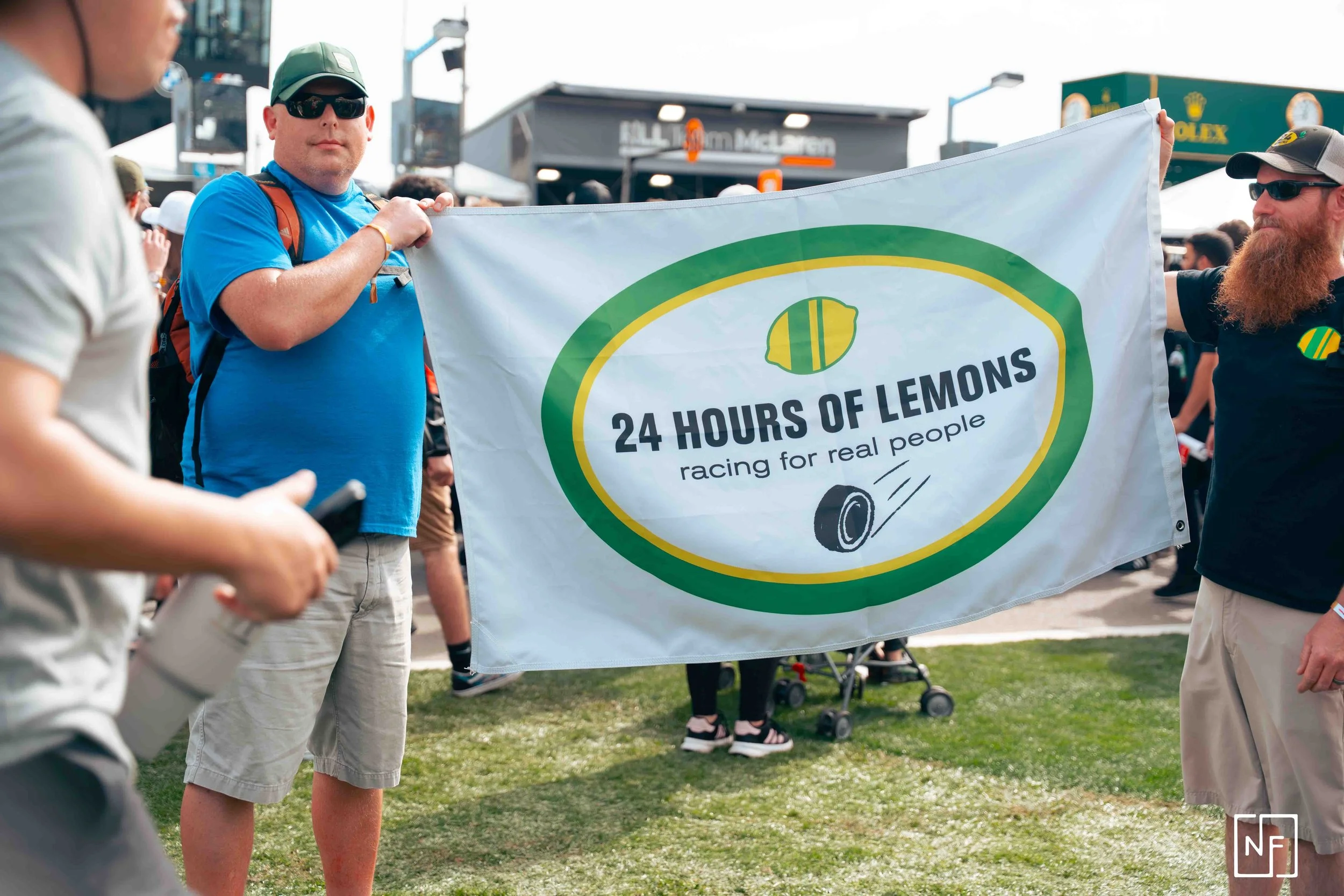 People holding a flag that reads '24 Hours of Lemons racing for real people' at an outdoor event.