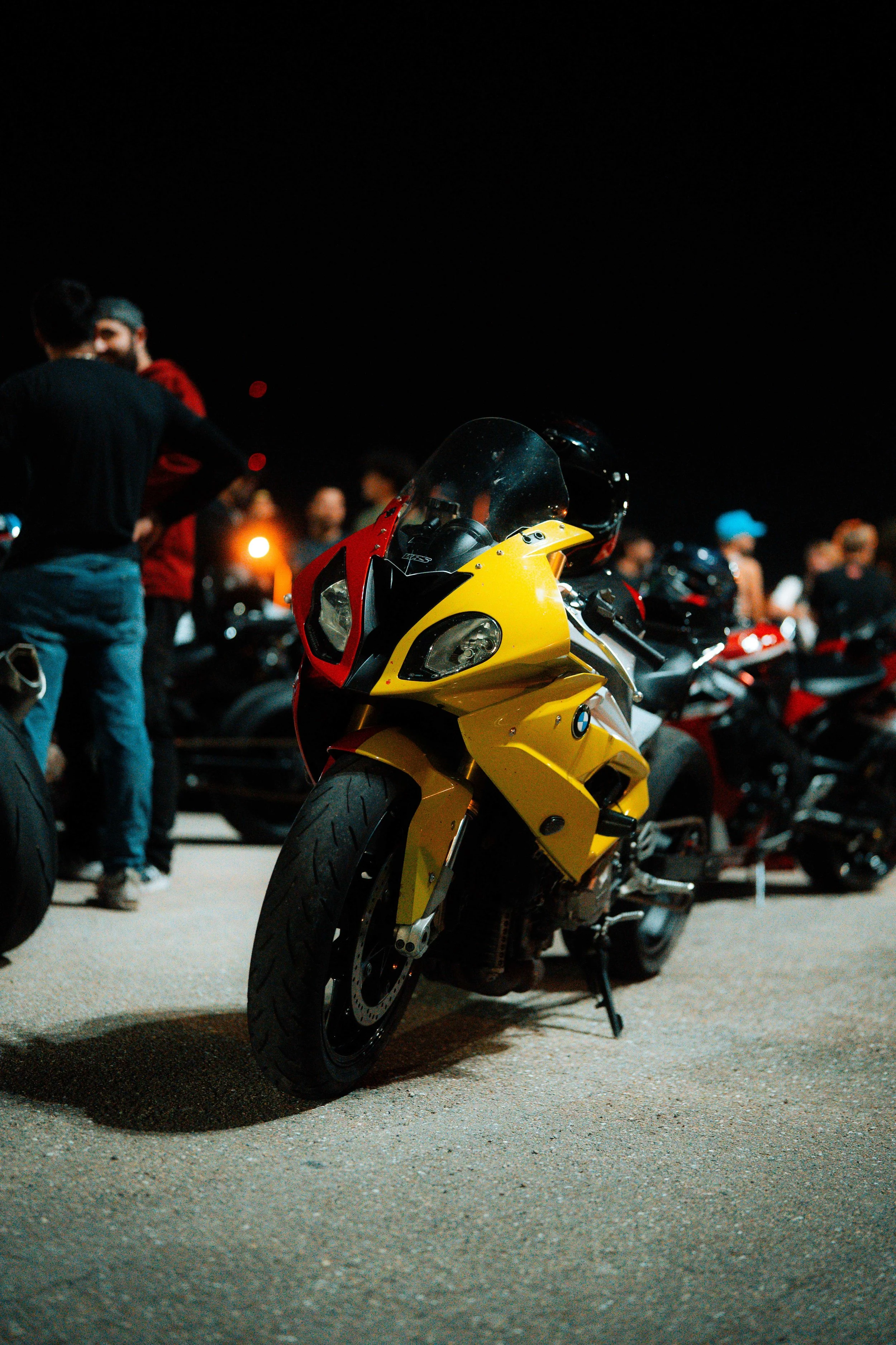 Yellow and red sport motorcycle parked among other bikes at night, with people gathered in the background.