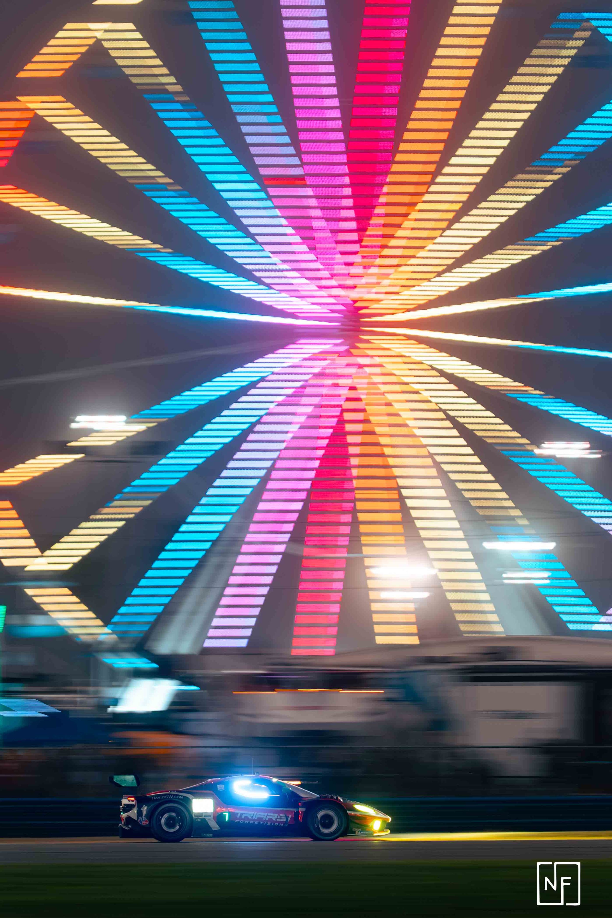 A race car with the number 7 and sponsor logos driving on a track at night, with colorful neon lights forming a dynamic, tunnel-like pattern overhead.