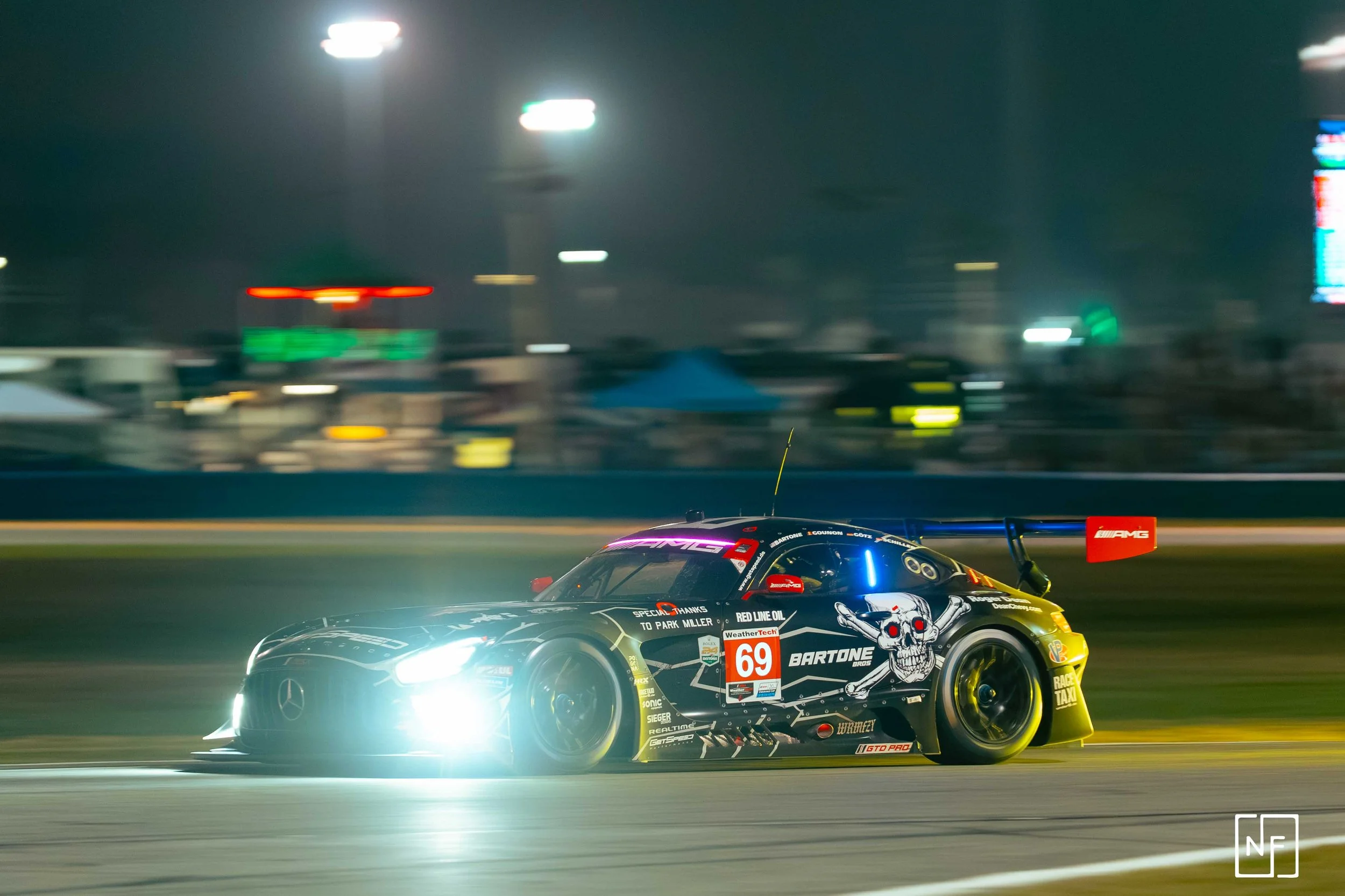 Nighttime race car speeding on track with bright headlights and motion blur, featuring a skull graphic on the side and sponsor logos.
