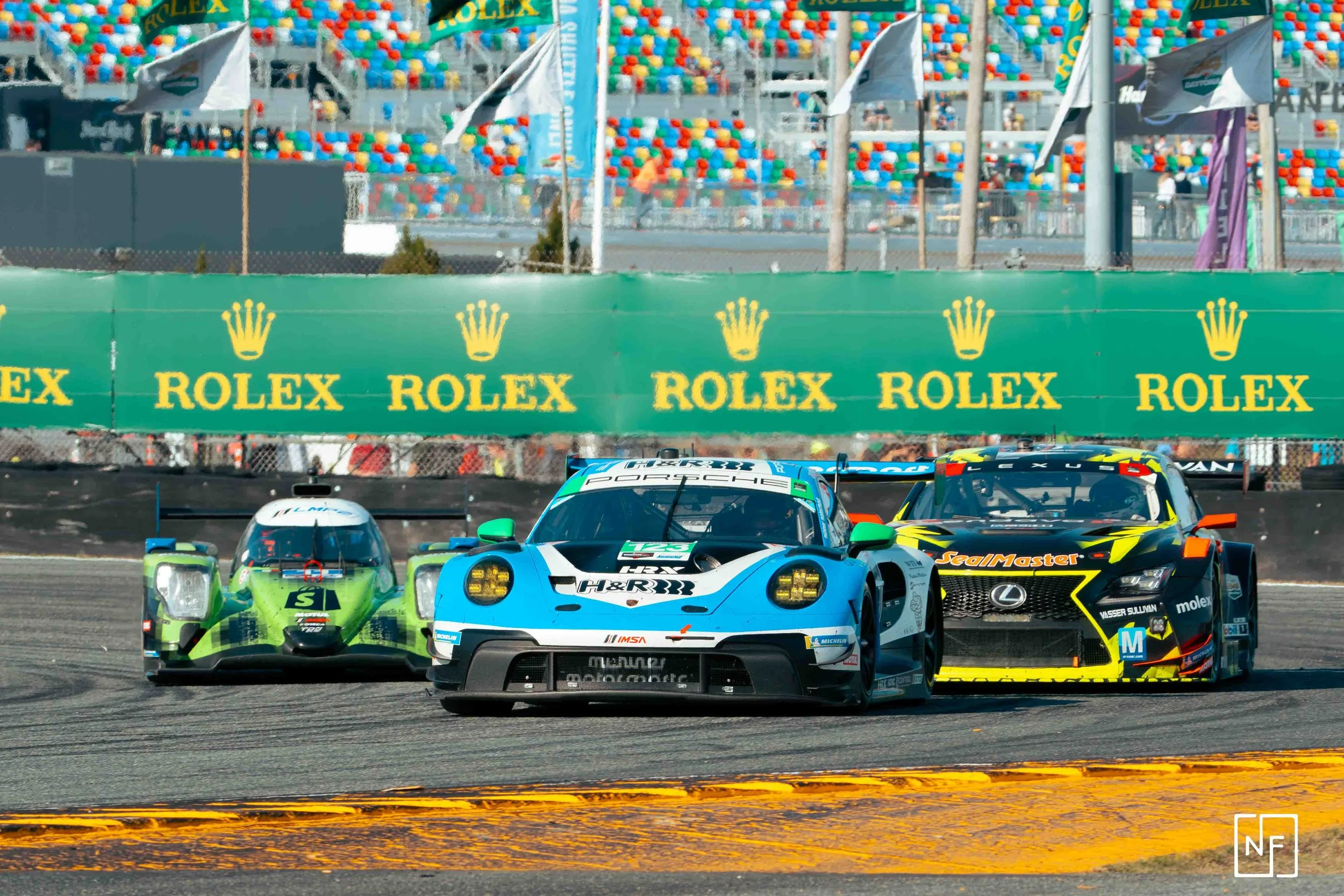 Three race cars on a track, with a green Rolex advertising banner and stands filled with colorful seats in the background.