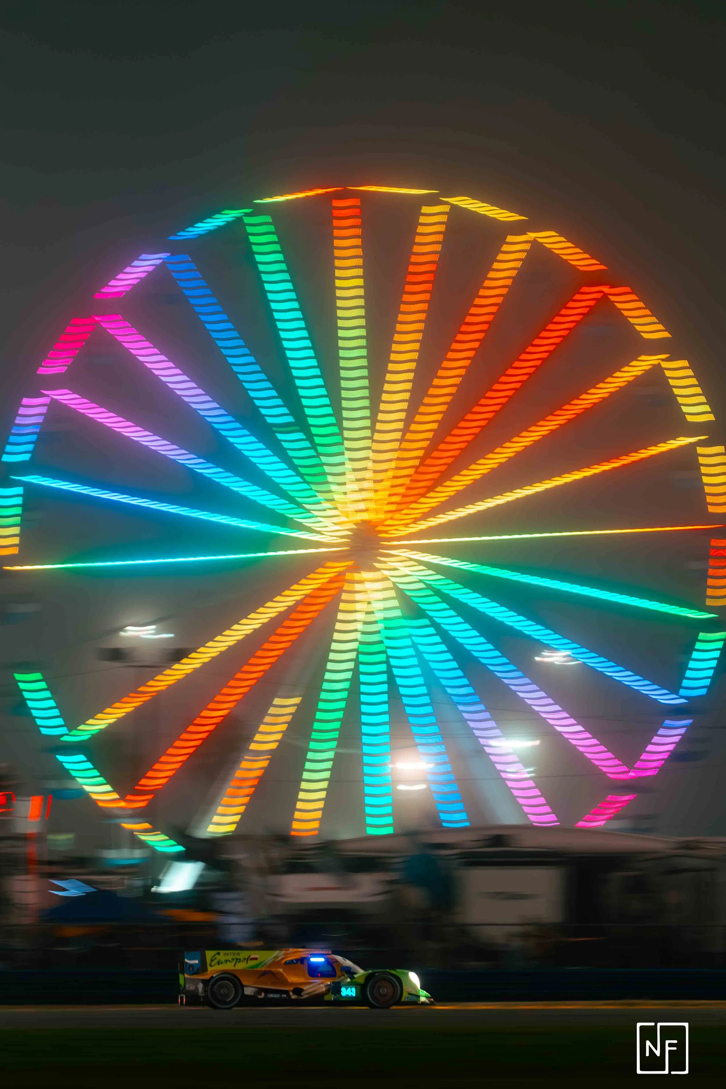 Colorful illuminated Ferris wheel at night with a race car passing in front.