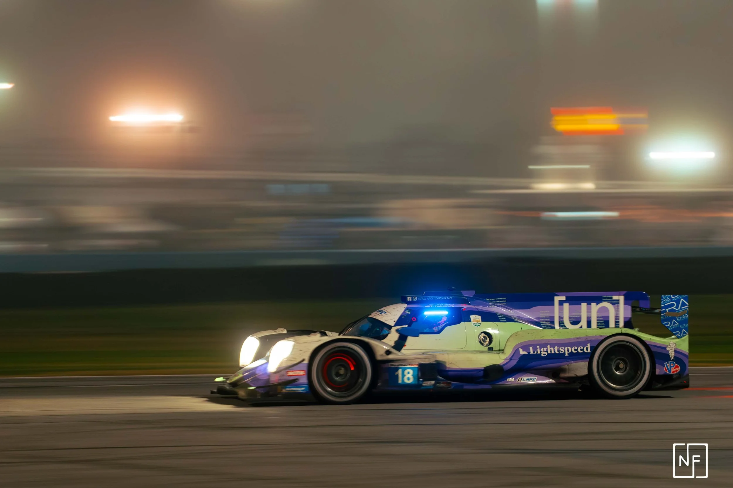 A racing car on a track at night with blurred lights in the background, illuminated headlights, and a blue light on top.