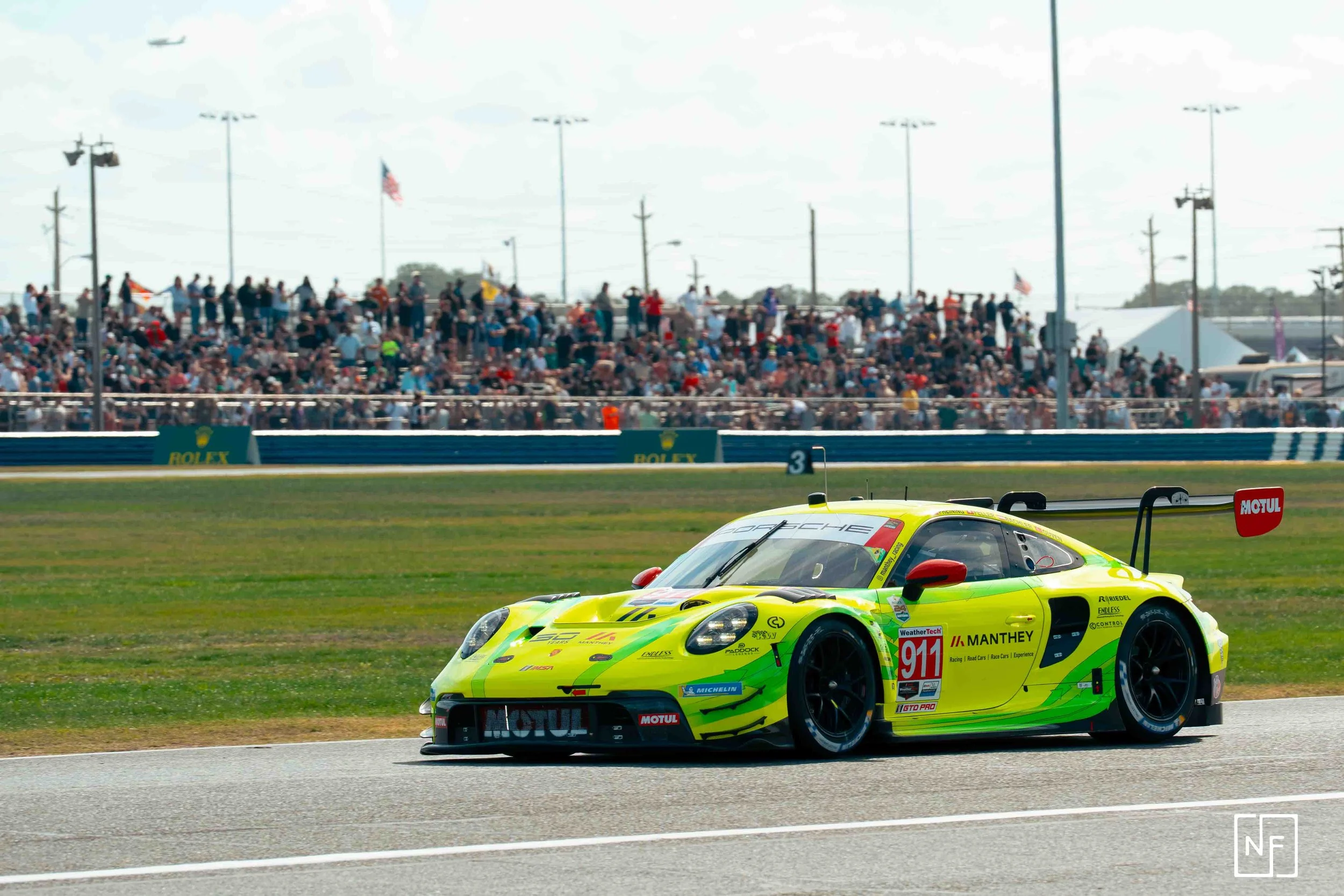 A neon yellow race car with the number 911 on it speedily driving on a race track during a car race, with a large crowd of spectators in the grandstand in the background.