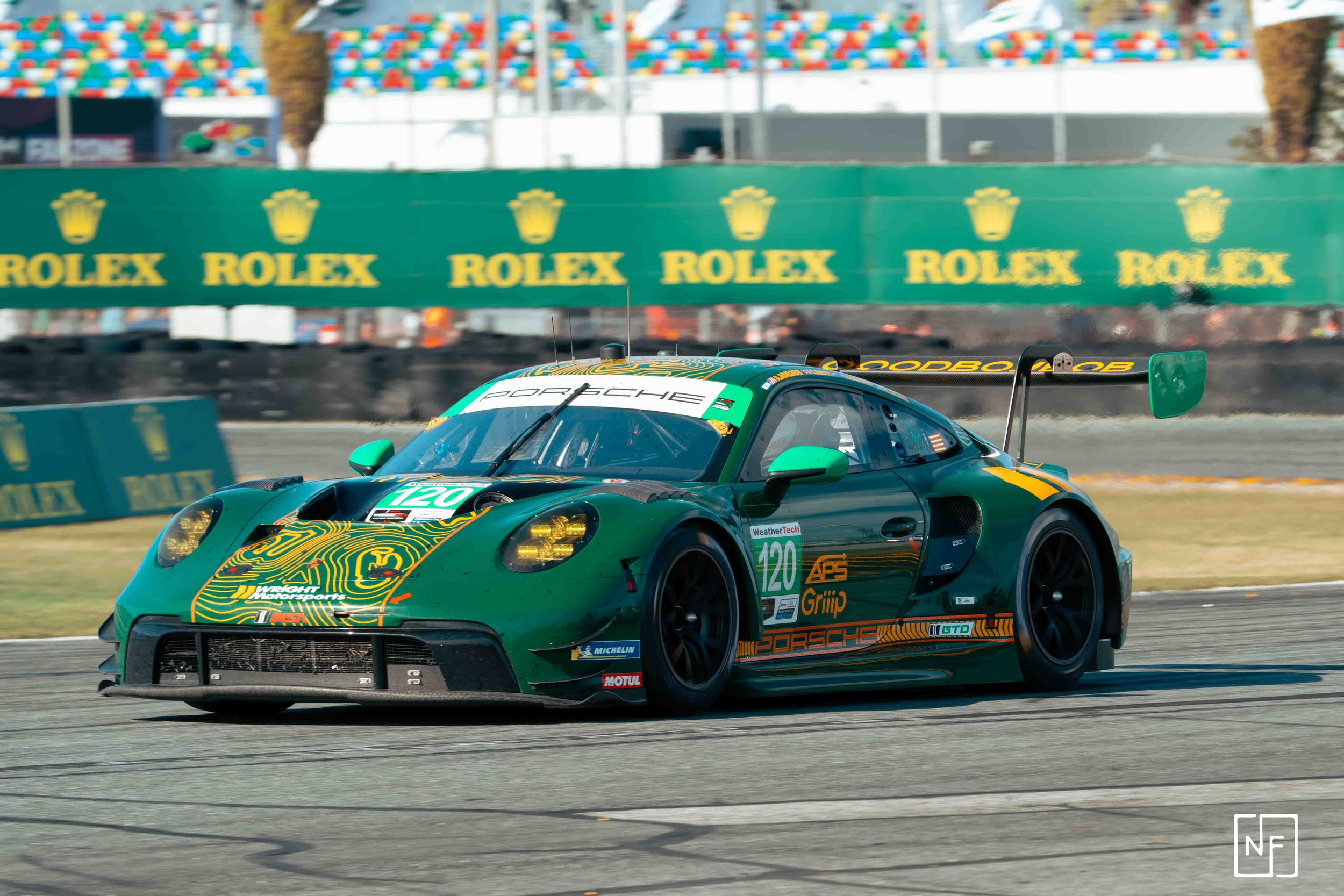 A green race car with black and yellow accents speeding on a race track. The car has sponsorship decals, including Porsche and Michelin. The background features green Rolex banners and colorful decorations.