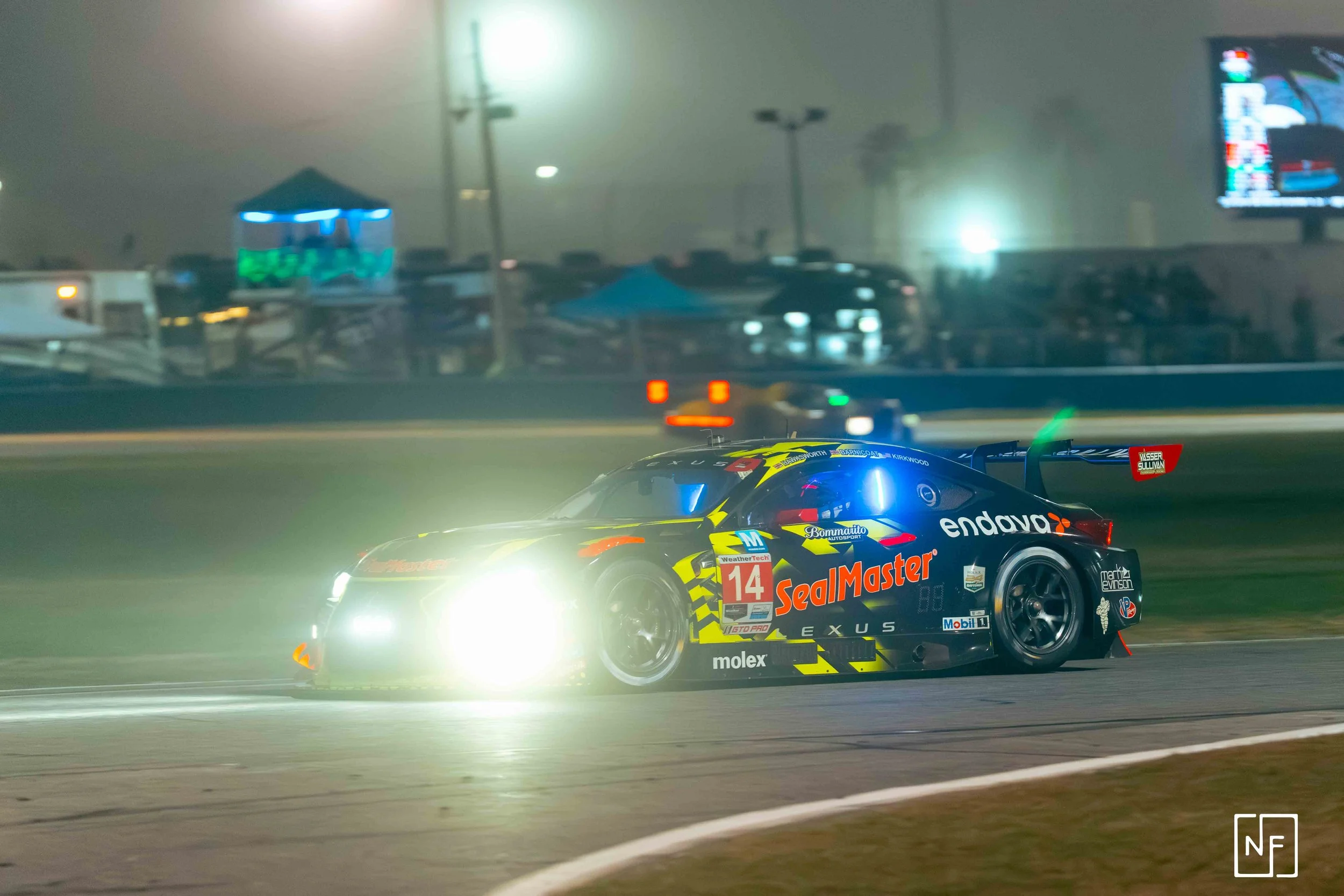 A race car with bright headlights speeding on a track at night, with illuminated scenery and blurred background.