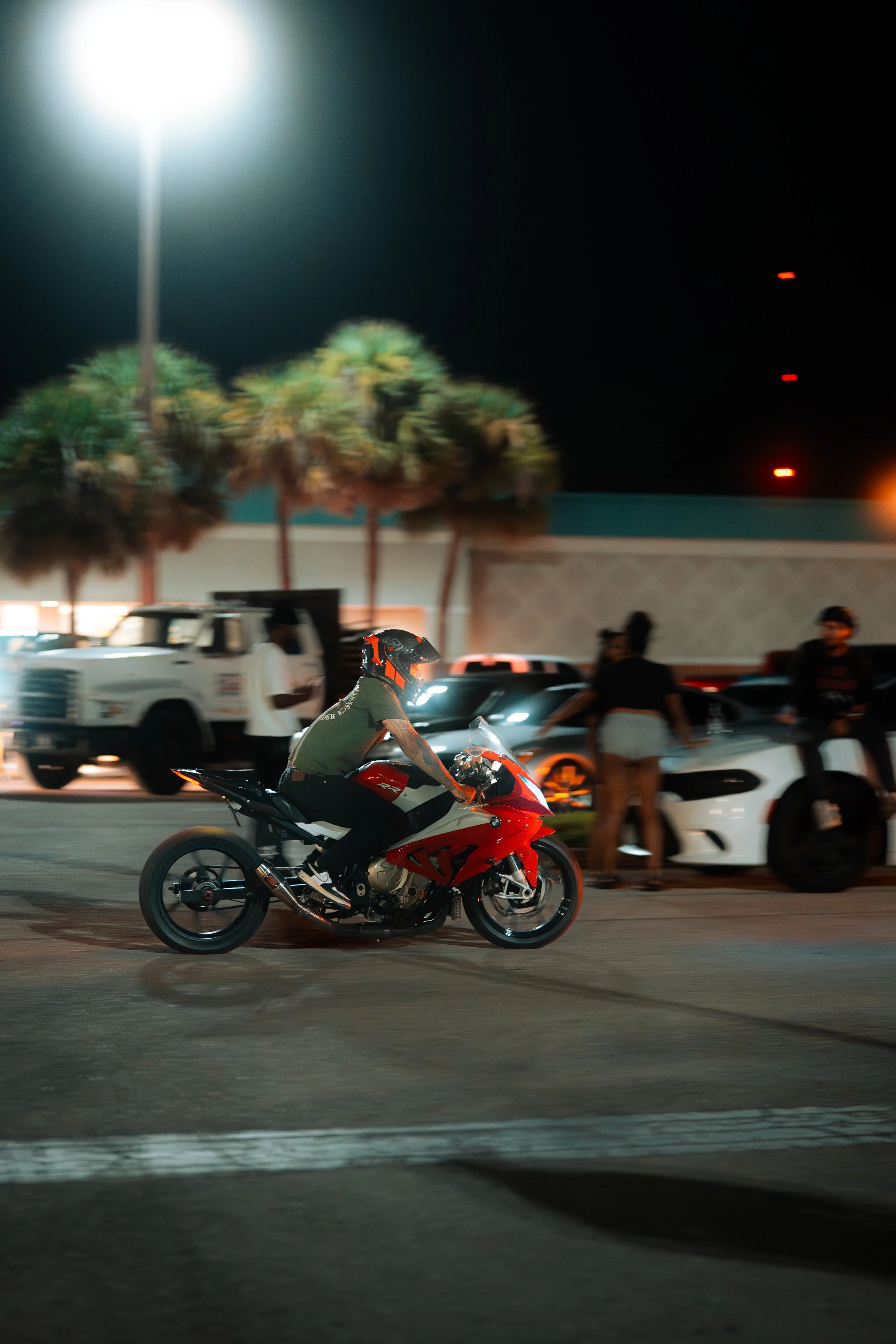 A motorcyclist wearing a helmet riding a red and white motorcycle on a city street at night, with cars and pedestrians in the background.