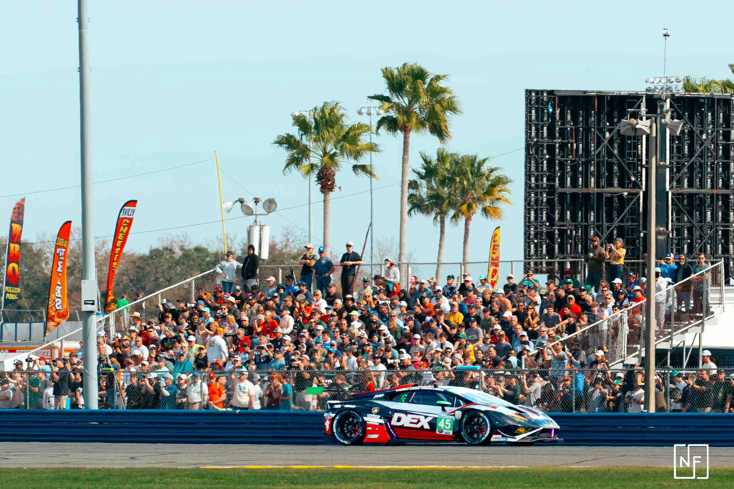 A race car with the number 45 on it driving on a racetrack, with a large crowd of spectators watching from the bleachers in the background, palm trees, and flags.