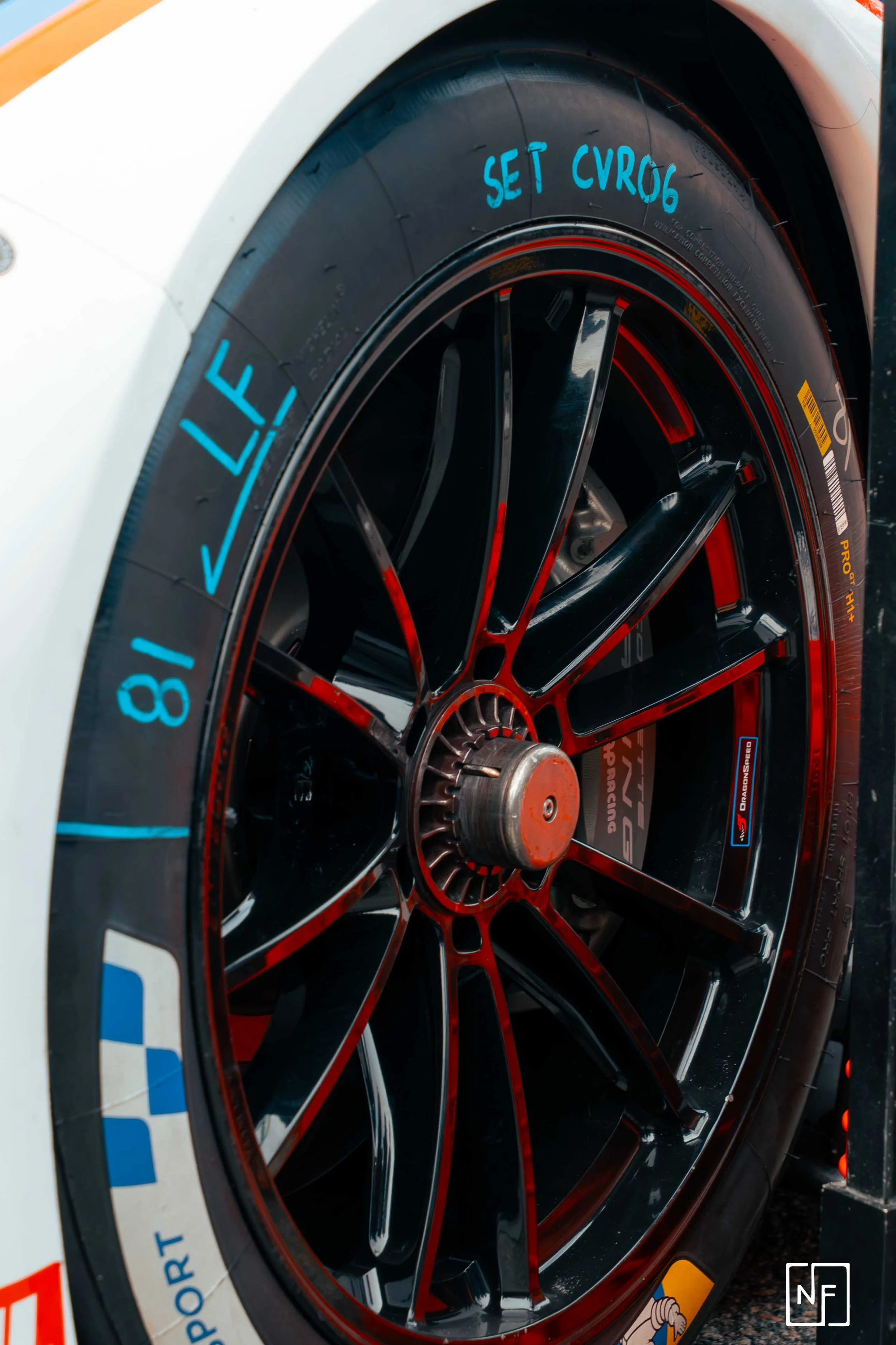 Close-up of a race car wheel with black and red rim and blue markings on the black tire, with writing indicating tire pressure and setup details.
