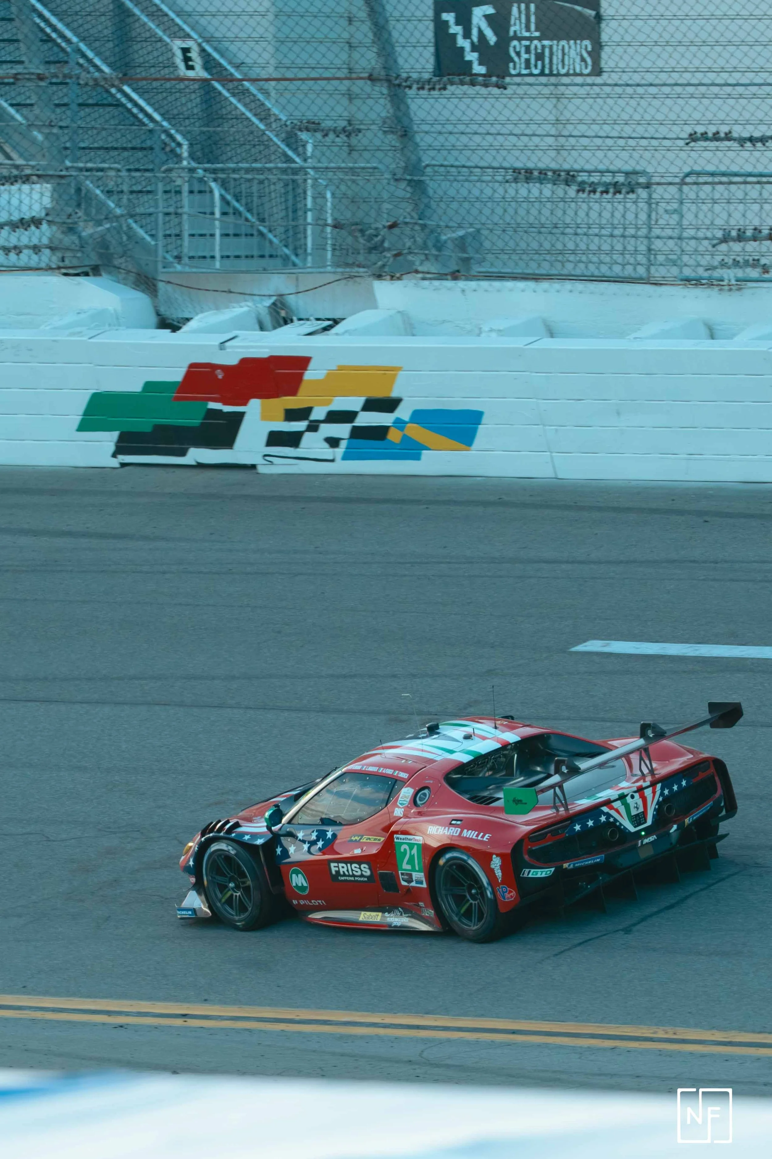 A red race car with patriotic decals on a racetrack with a 'All Sections' sign and a colorful painted wall in the background.