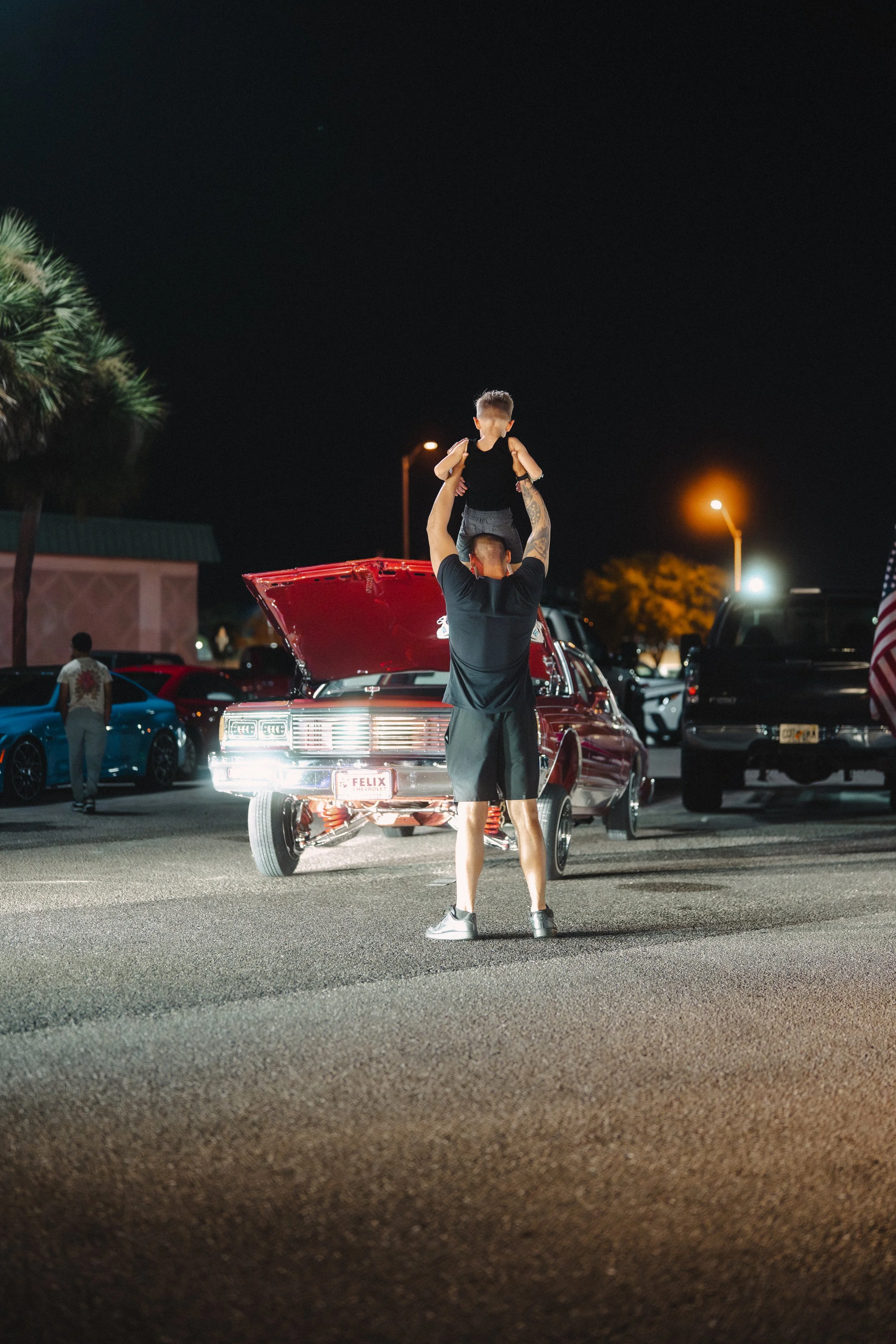 A man lifting a child in the air near a red car with its hood open at night in a parking lot.
