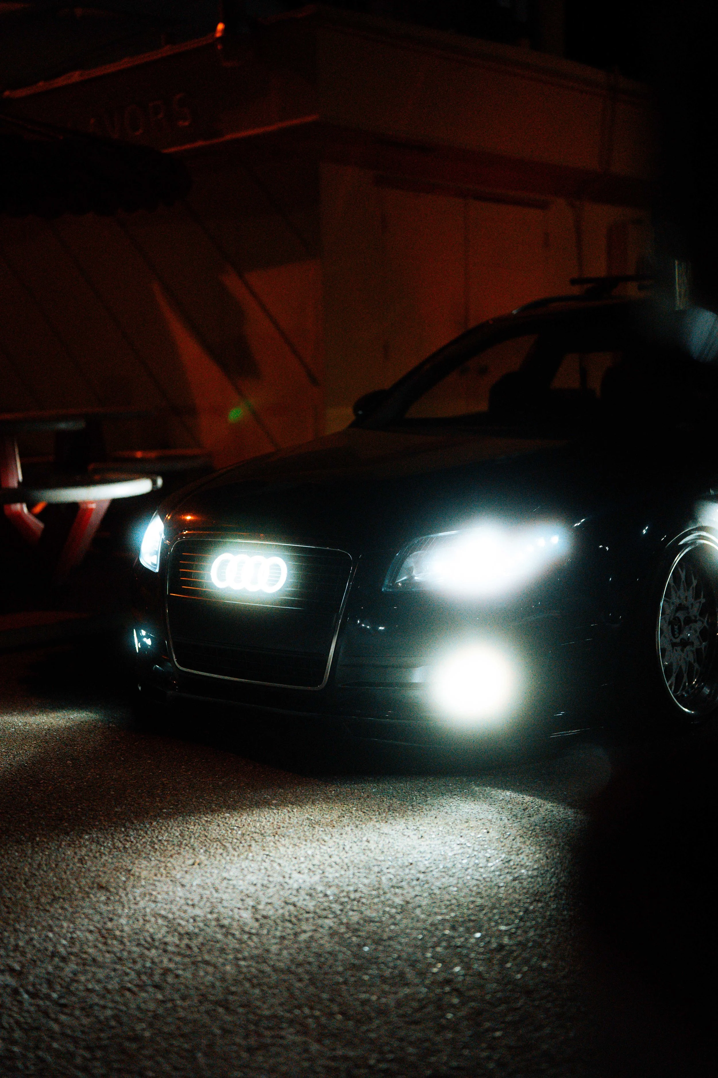 A black Audi car with its headlights and grille illuminated, parked on a textured asphalt surface at night. The front license plate is not visible, and there is a building with a sign that reads 'NORS' in the background.