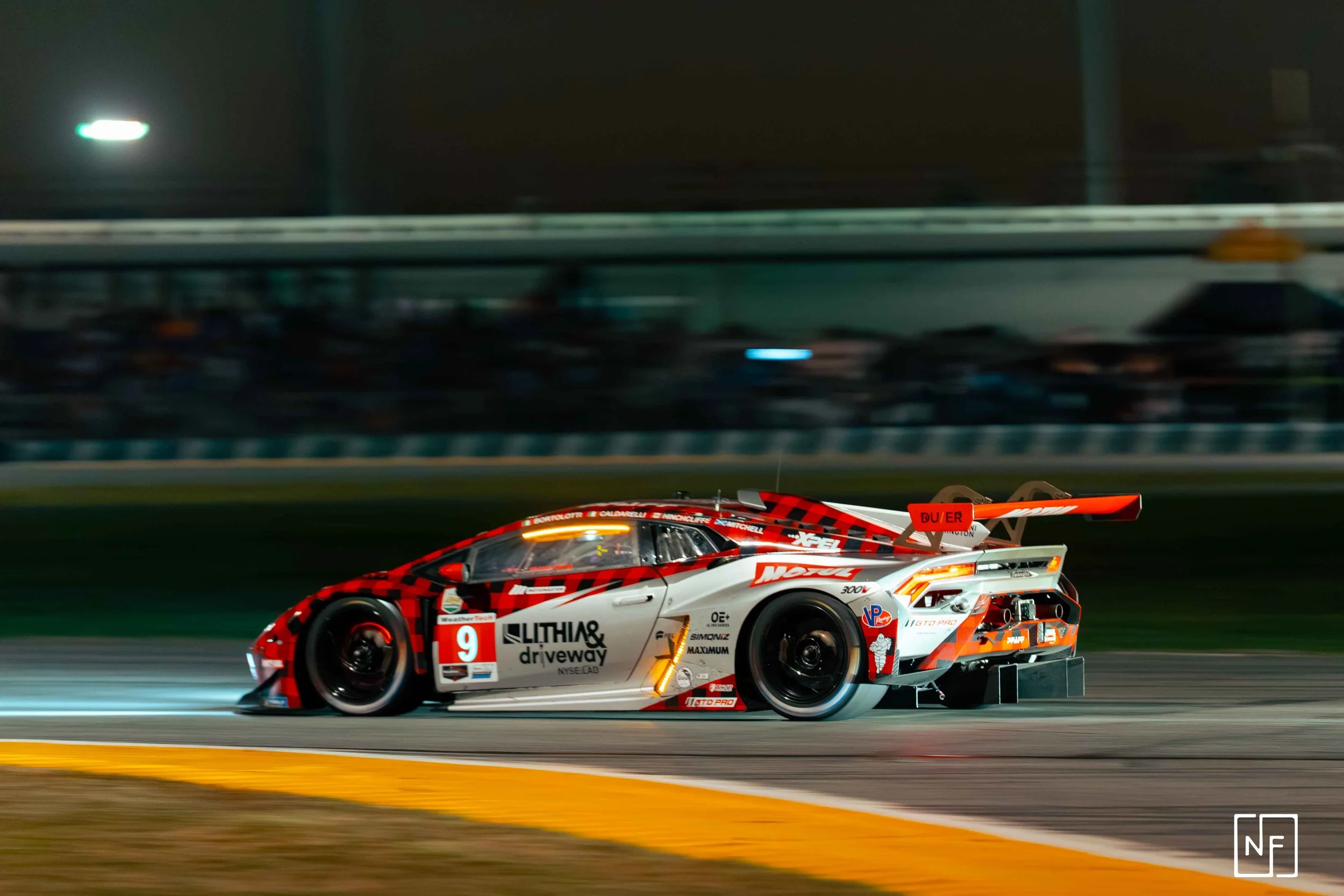 Race car with red, white, and black livery speeding on a race track at night, blurred spectators in background.
