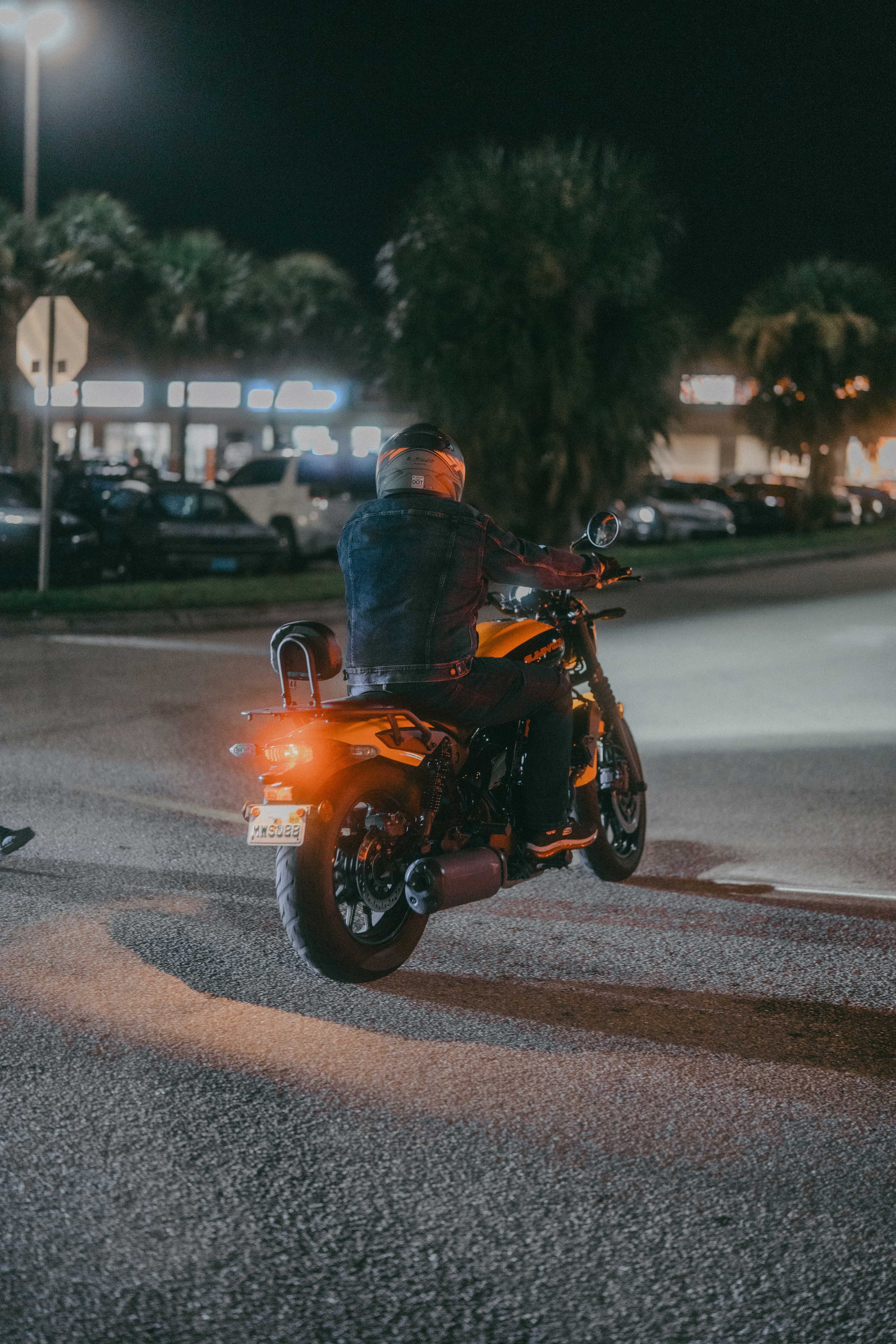 A person riding a motorcycle at night on a well-lit street, with parked cars and trees in the background.