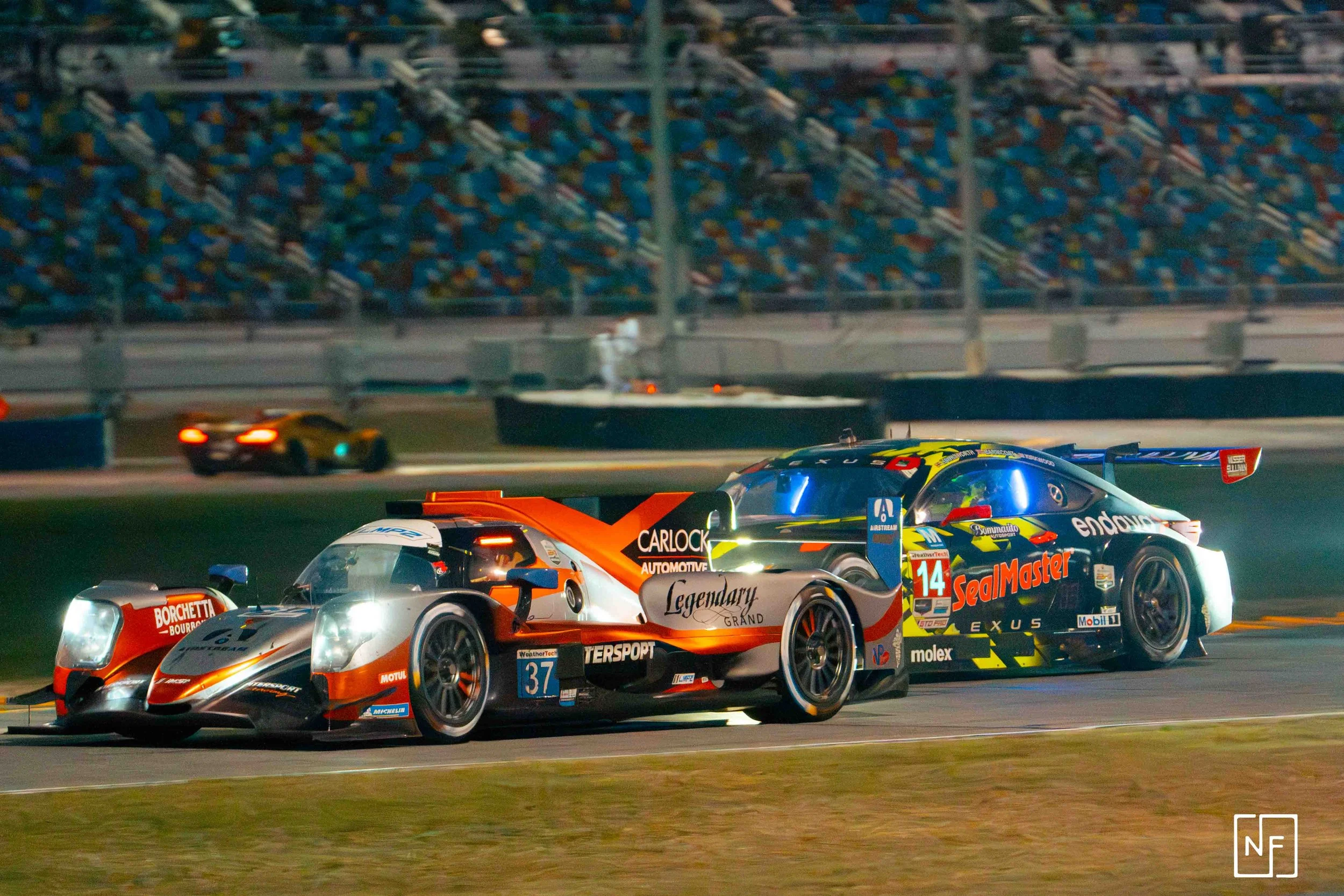 A race car with orange and black colors and the number 37, and a dark blue and yellow race car with the number 14, competing on a nighttime race track.