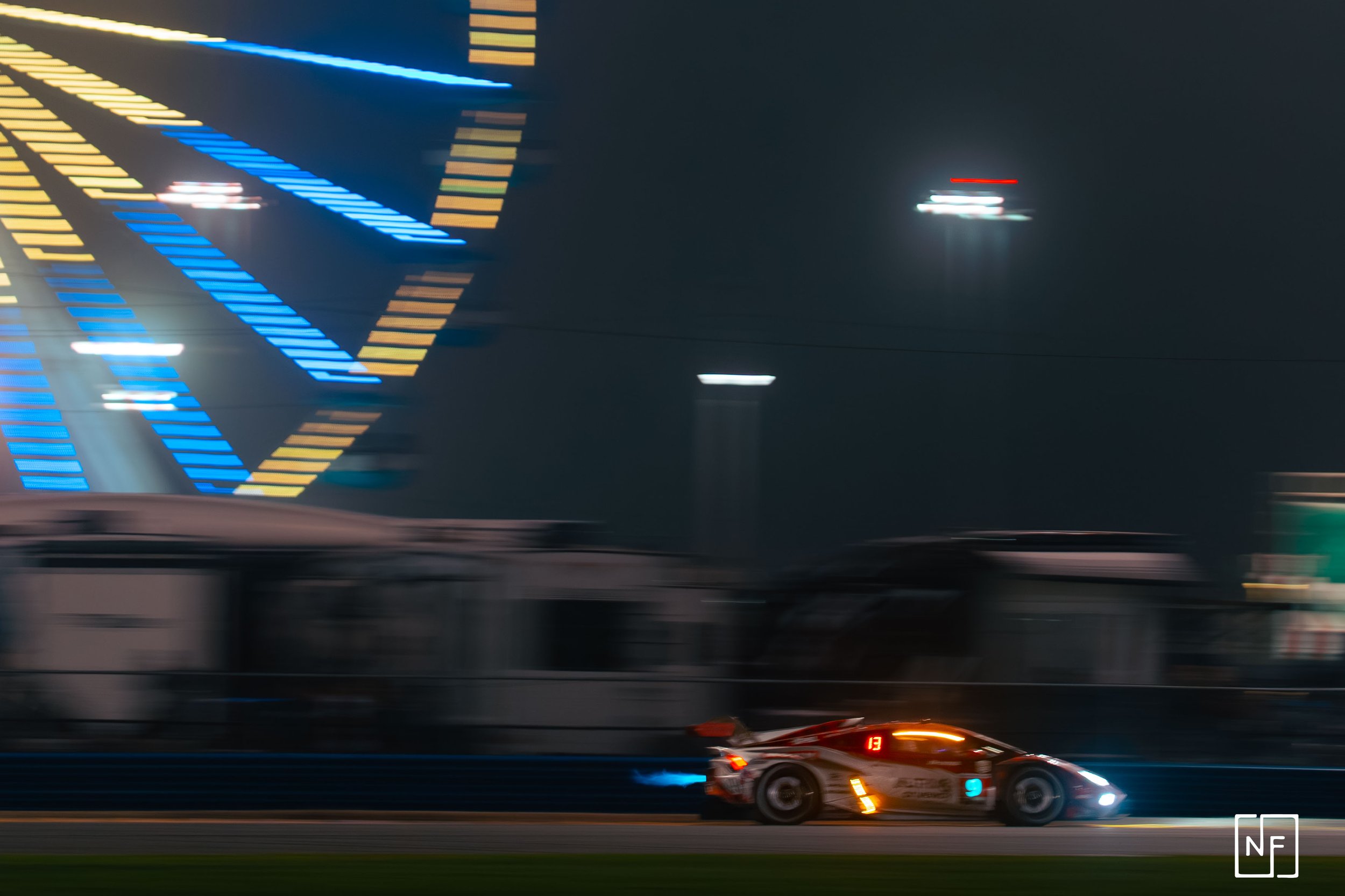 A race car moving at high speed on a racetrack at night, with blurred background and illuminated digital displays on a large screen in the background.