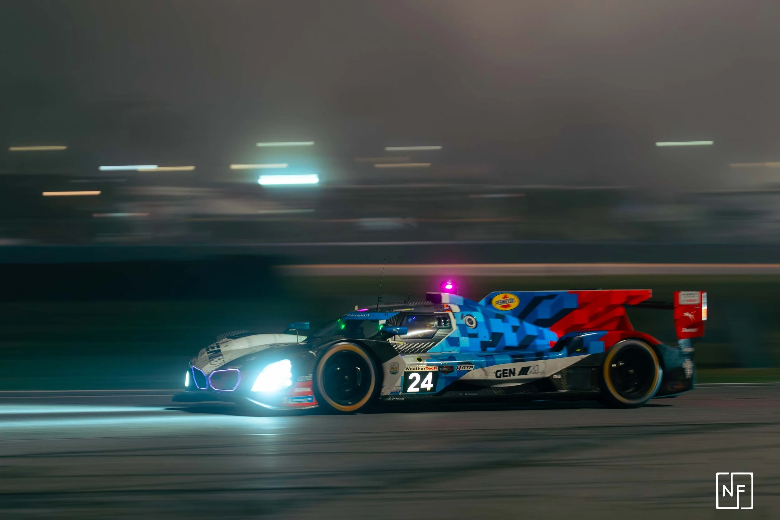 Nighttime image of a race car with headlights on, moving at high speed on a track, with blurred background lights.