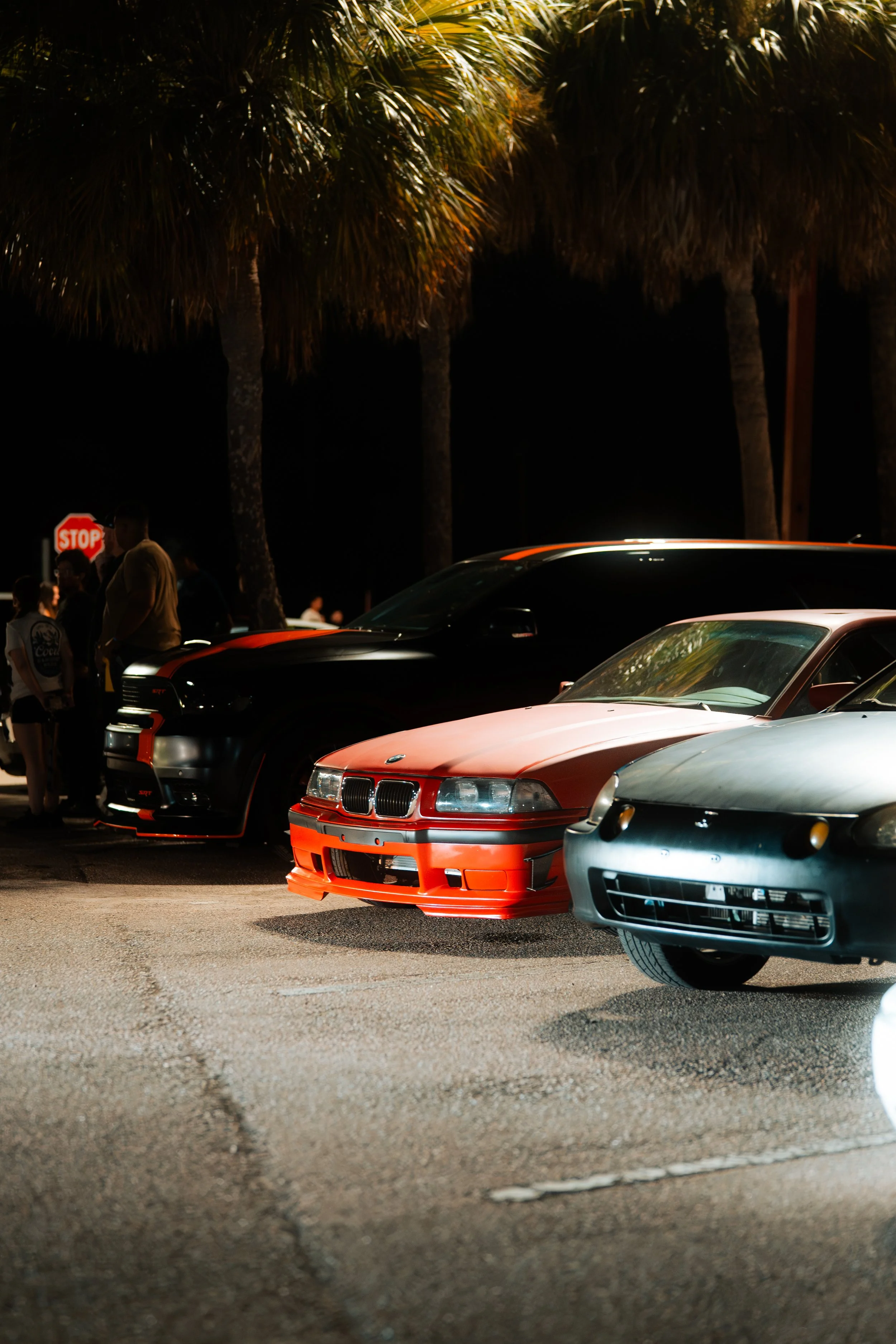 A nighttime scene with a row of parked cars, including a silver sports car, a red car, and a black vehicle, with palm trees and a stop sign in the background, and people gathered near the cars.