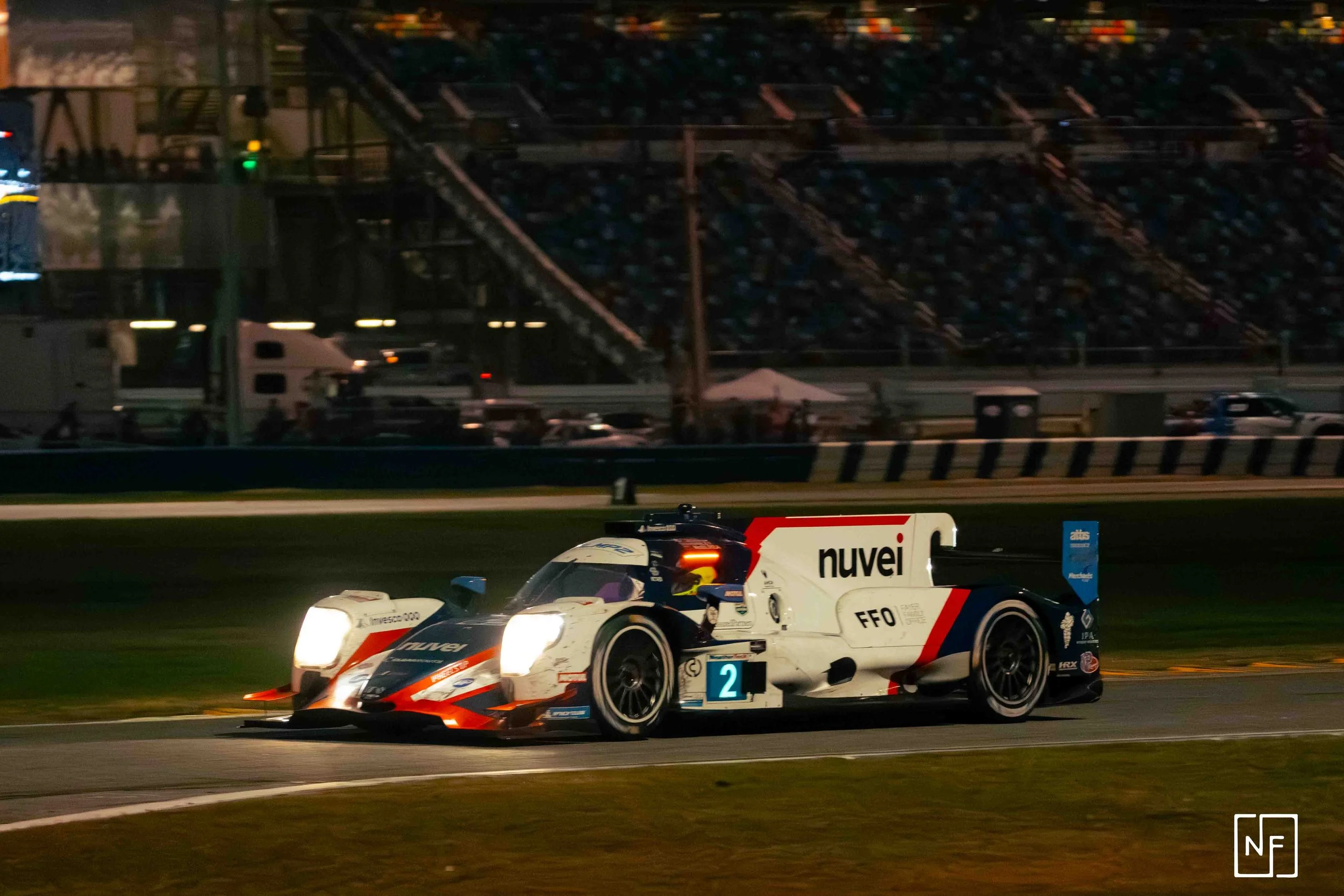 A race car driving on a track at night, illuminated by headlights, with audience stands in the background.