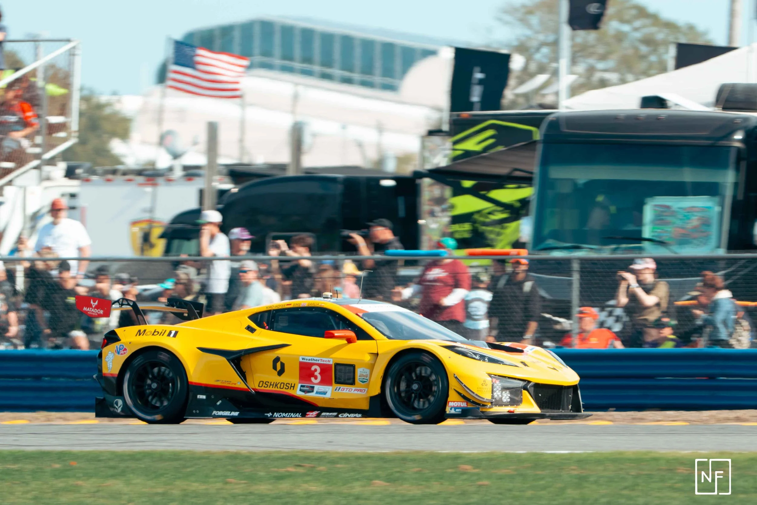 Yellow race car speeding on track with spectators watching in the background.