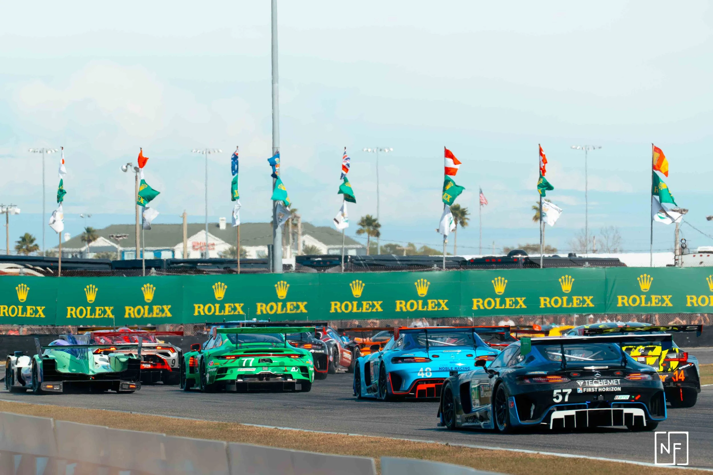 Multiple race cars on a track, competing in a race, with flags and advertising banners in the background.