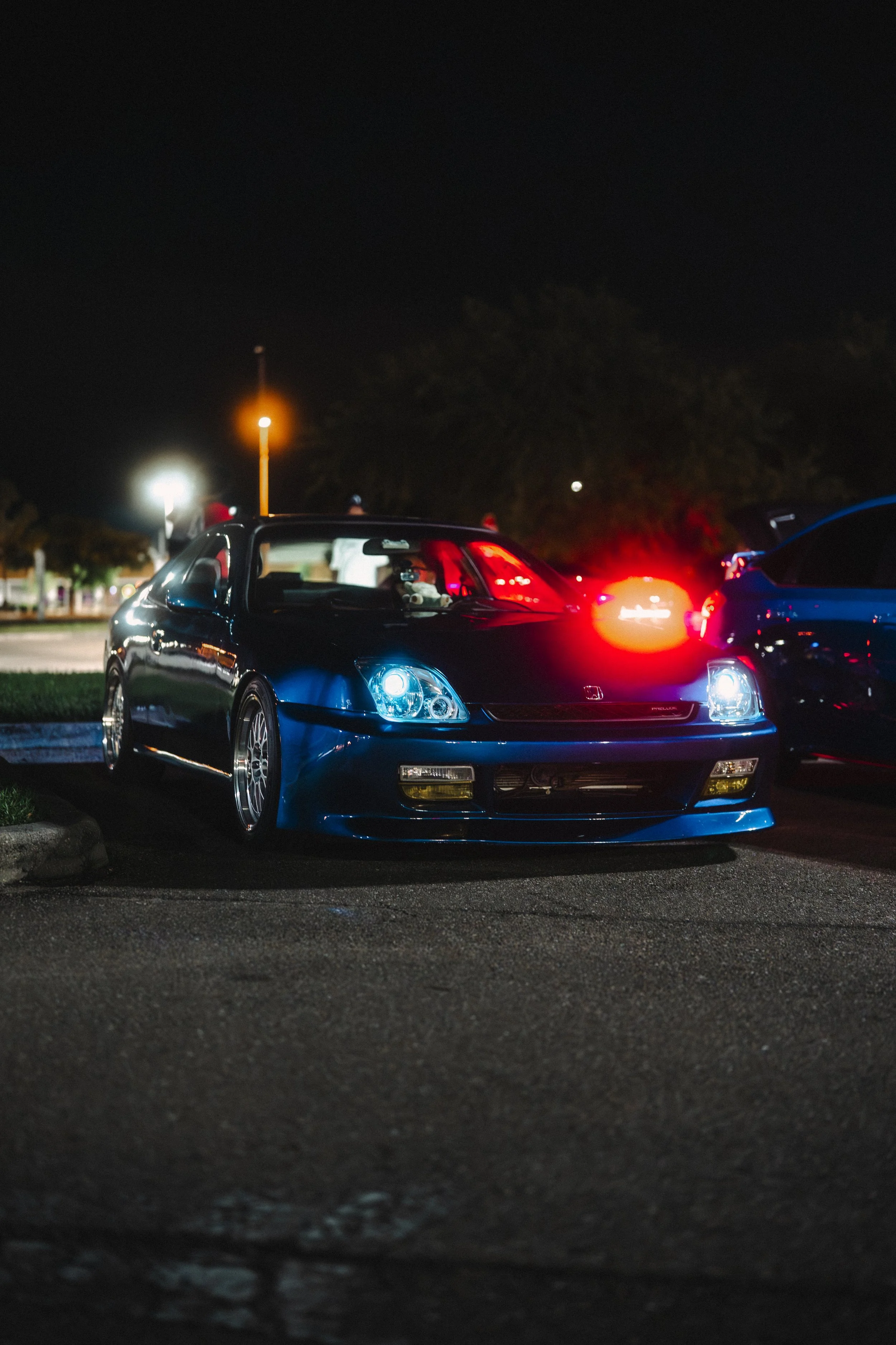 A black sports car parked at night in a parking lot with illuminated headlights and a red taillight. There are other cars and streetlights in the background.