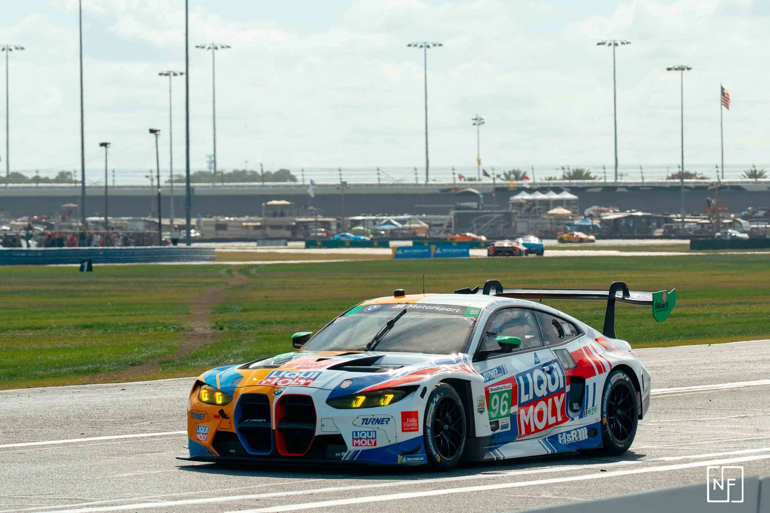 A race car on a track with a crowd and tents in the background, under a cloudy sky.